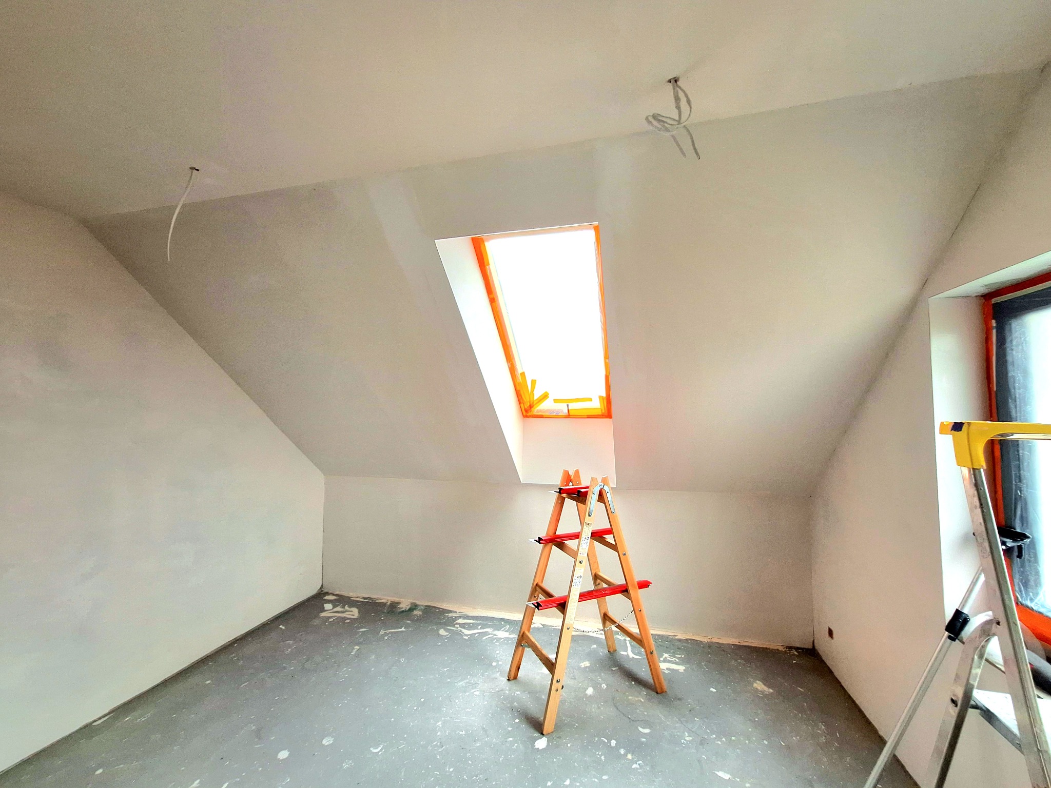Poddasze w trakcie wykańczania, widoczna drewniana drabina, ślady po gładzi na ścianach i podłodze, okno dachowe z pomarańczową ramą i wystające kable ze stropu.