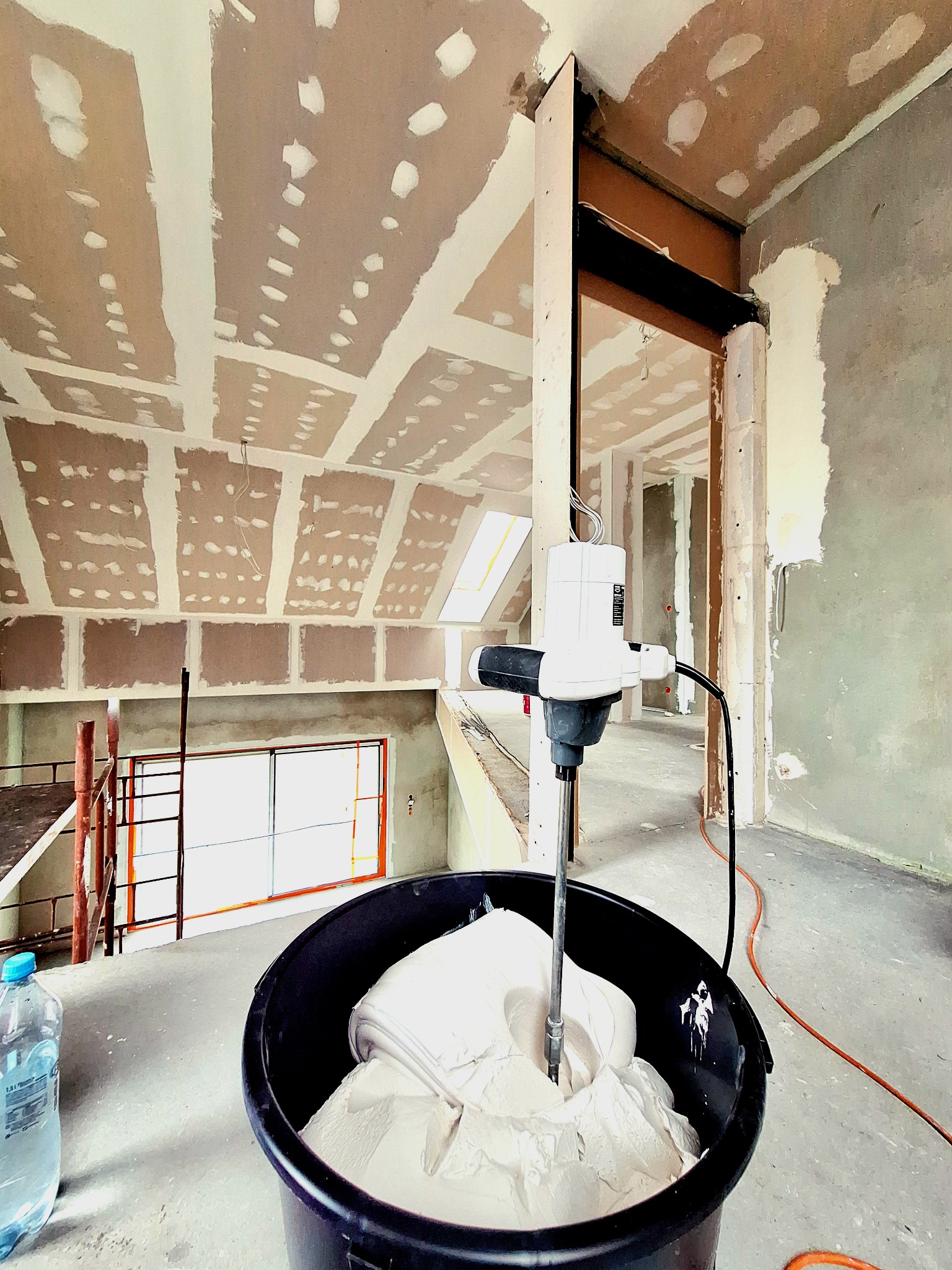 Mieszarka elektryczna w czarnym wiadrze z gładzią, w tle surowy strop z płyt kartonowo-gipsowych przed szpachlowaniem, widoczne rusztowanie i niedokończone ściany.