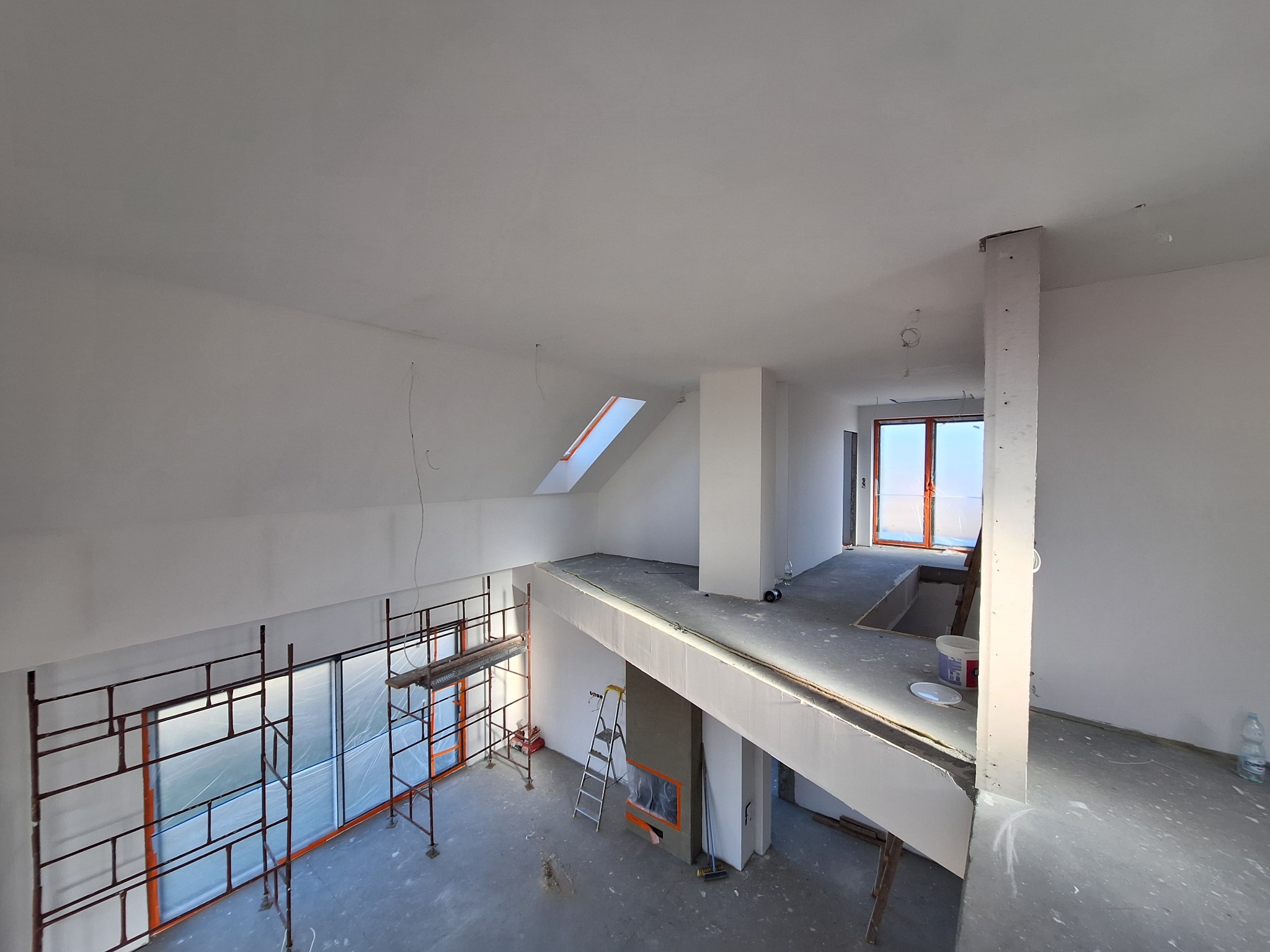 Przestronne wnętrze w trakcie prac wykończeniowych, widok z góry na antresolę, rusztowania przy oknach, świeżo pomalowane ściany na biało, widoczne ślady po gładzi, drabina oparta o kominek.