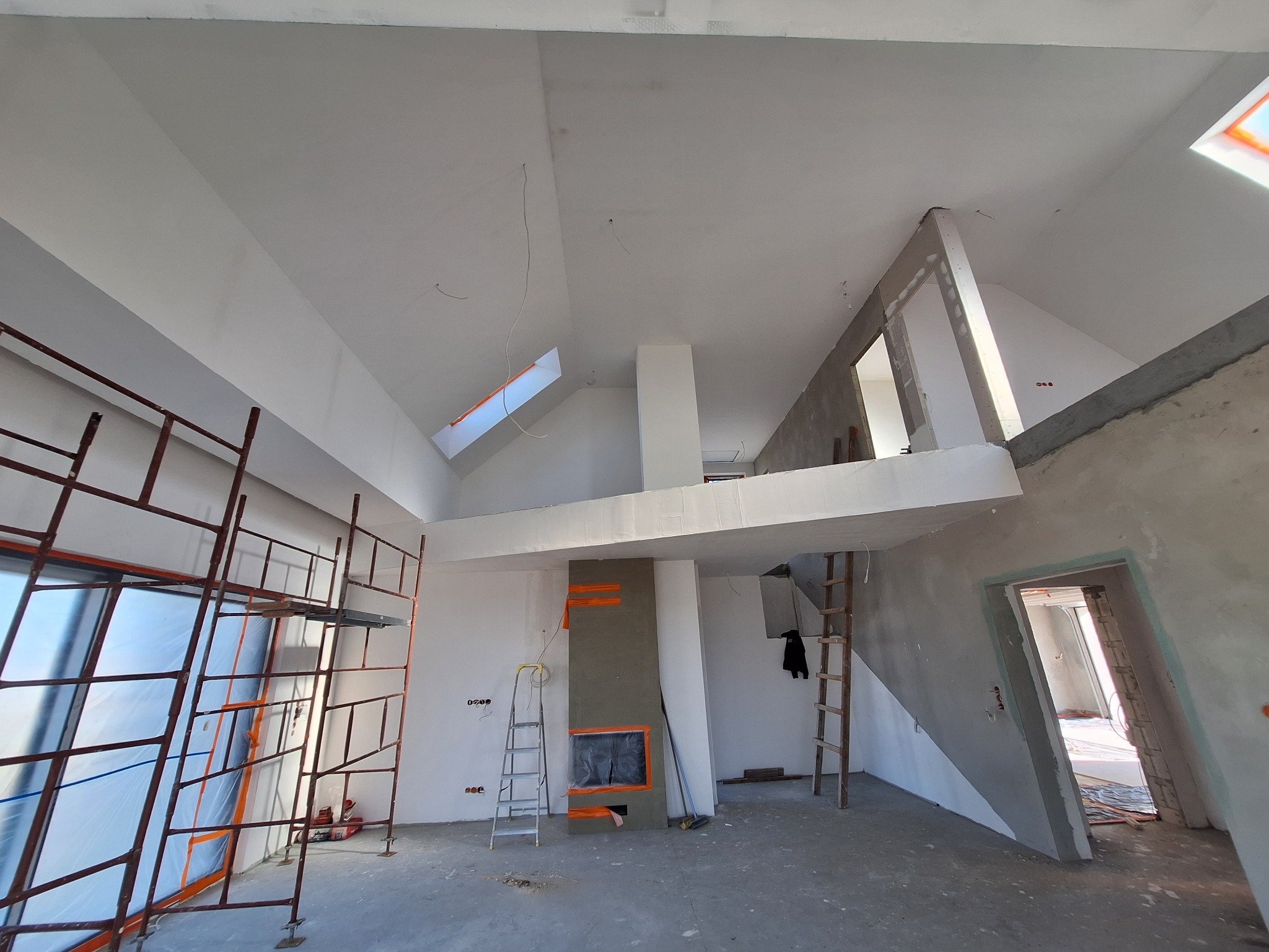 Wnętrze w trakcie prac wykończeniowych: widoczne rusztowania przy oknie z folią ochronną, ściany w stanie surowym lub po nałożeniu gładzi, przygotowany kominek z taśmą malarską, drabina oparta...