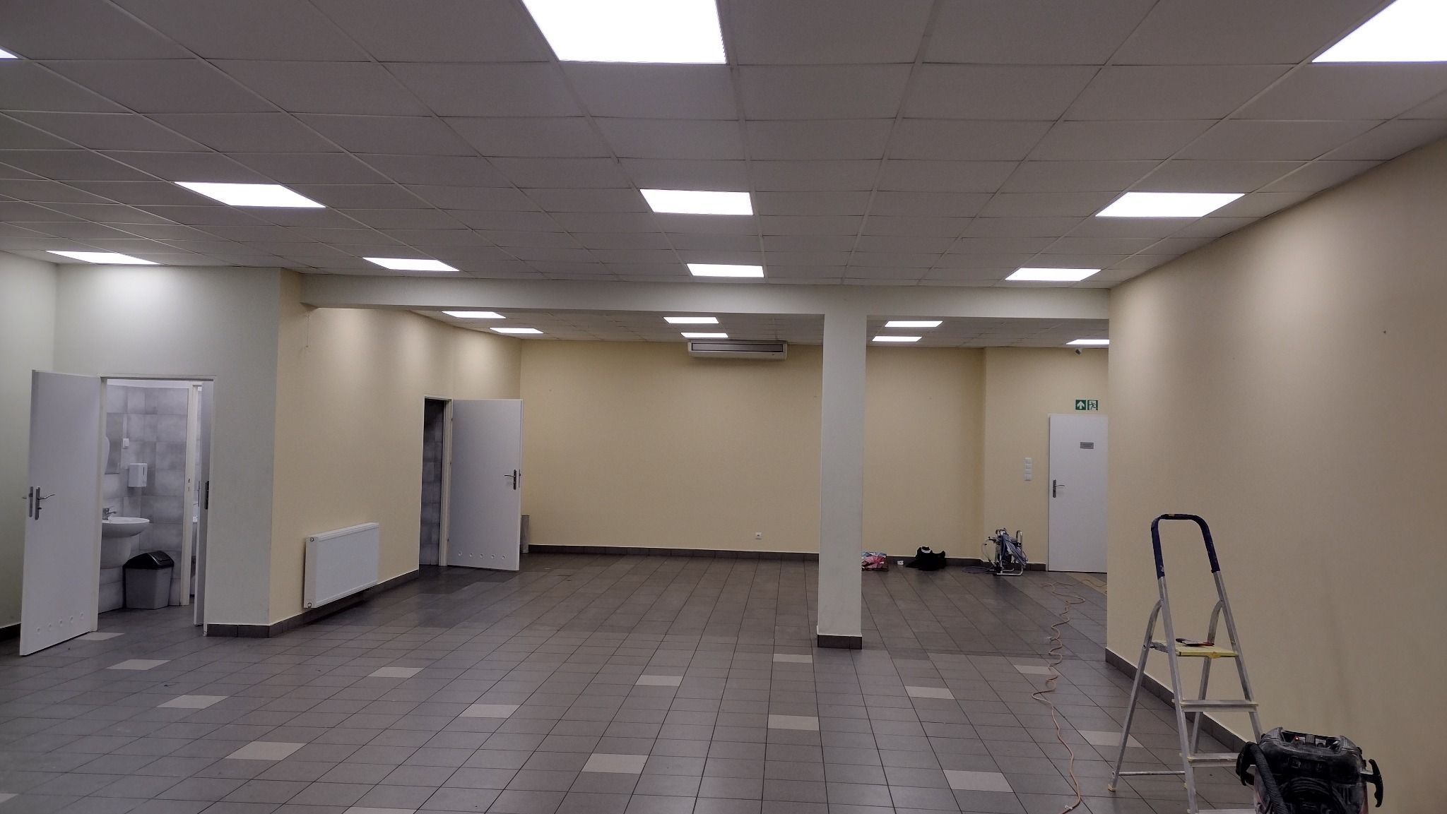 Przestronne wnętrze komercyjne po odświeżeniu ścian, z widoczną drabiną i urządzeniem malarskim, sugerującym prace wykończeniowe.