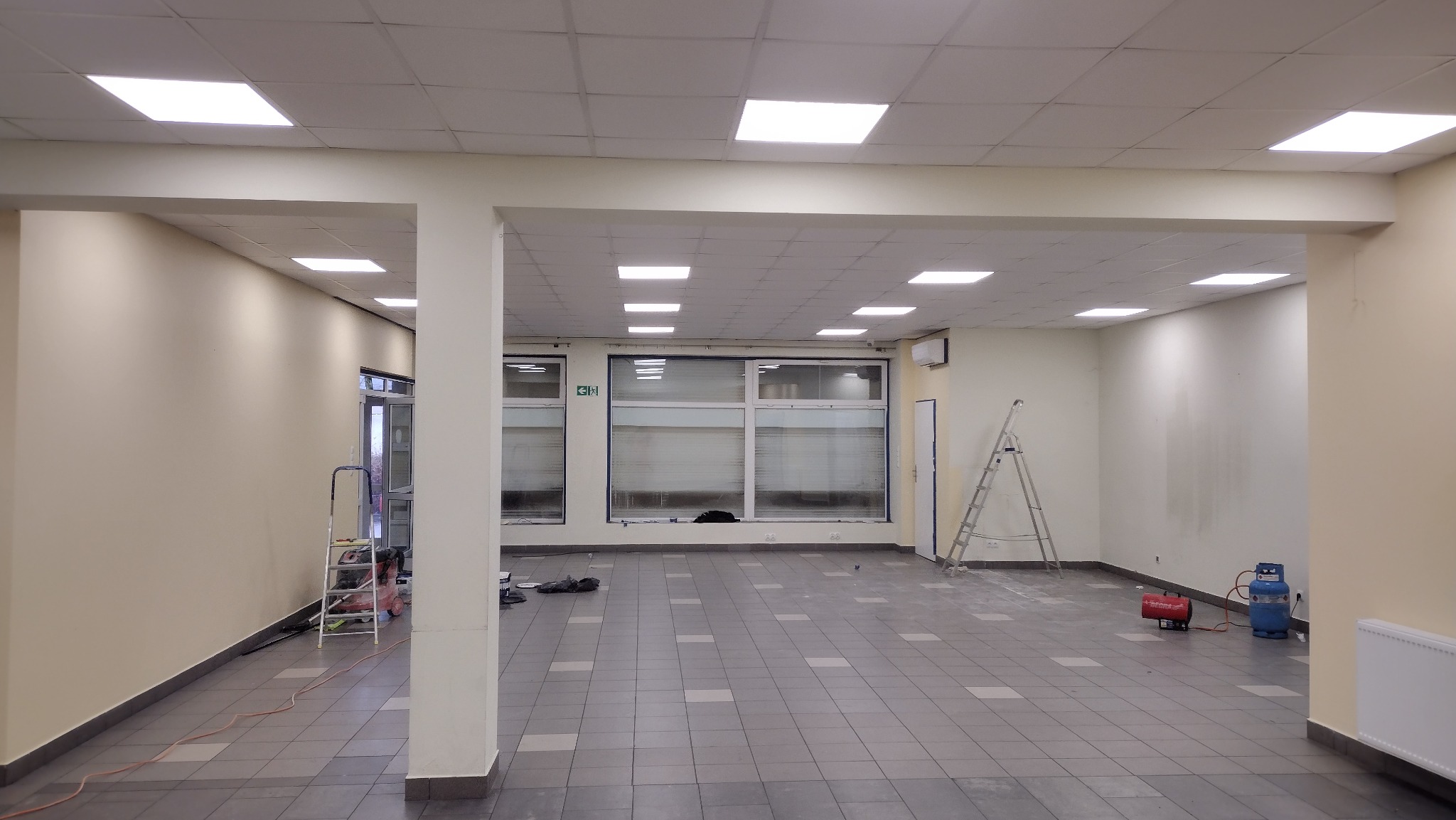 Duże, puste pomieszczenie biurowe w trakcie odświeżania, widoczne ślady po pracach, drabiny, grzejnik gazowy i pomalowane na jasny kolor ściany.