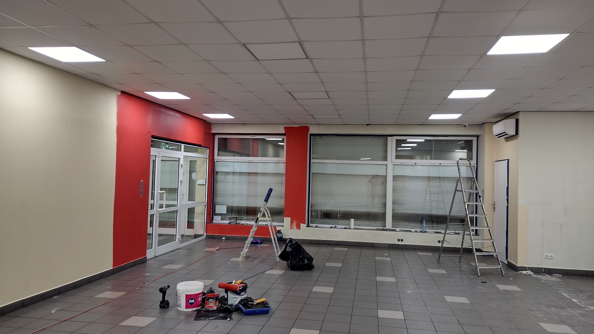 Malowanie ścian w biurze, widoczne fragmenty pomalowane na czerwono, drabiny, wiadro z farbą Atlas i Dulux oraz narzędzia malarskie na podłodze w szaro-białe kwadraty.