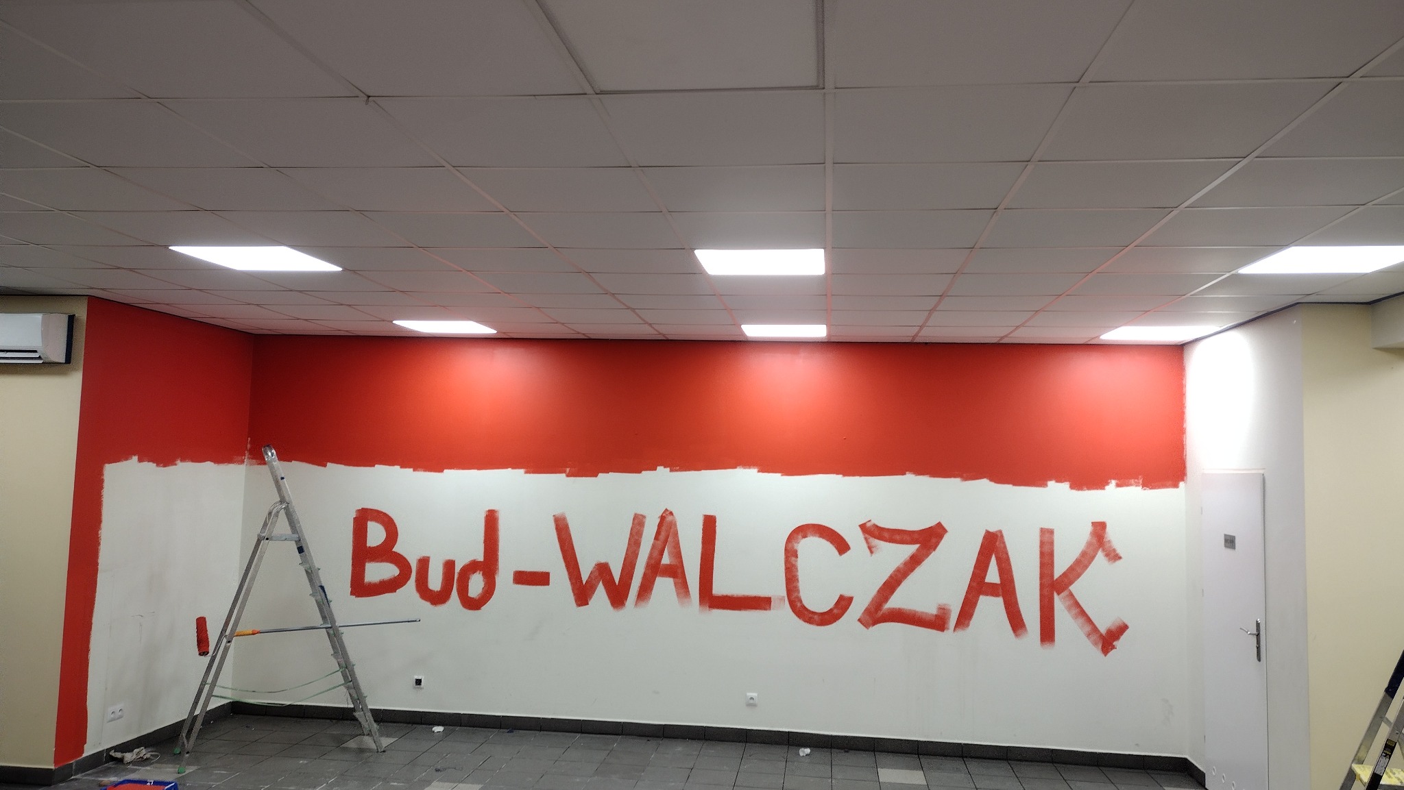 Świeżo pomalowana na czerwono górna część ściany w pomieszczeniu biurowym, z niepomalowanym pasem poniżej, widoczna drabina i wałek malarski, na ścianie napis 'Bud-WALCZAK' wykonany czerwoną farbą.