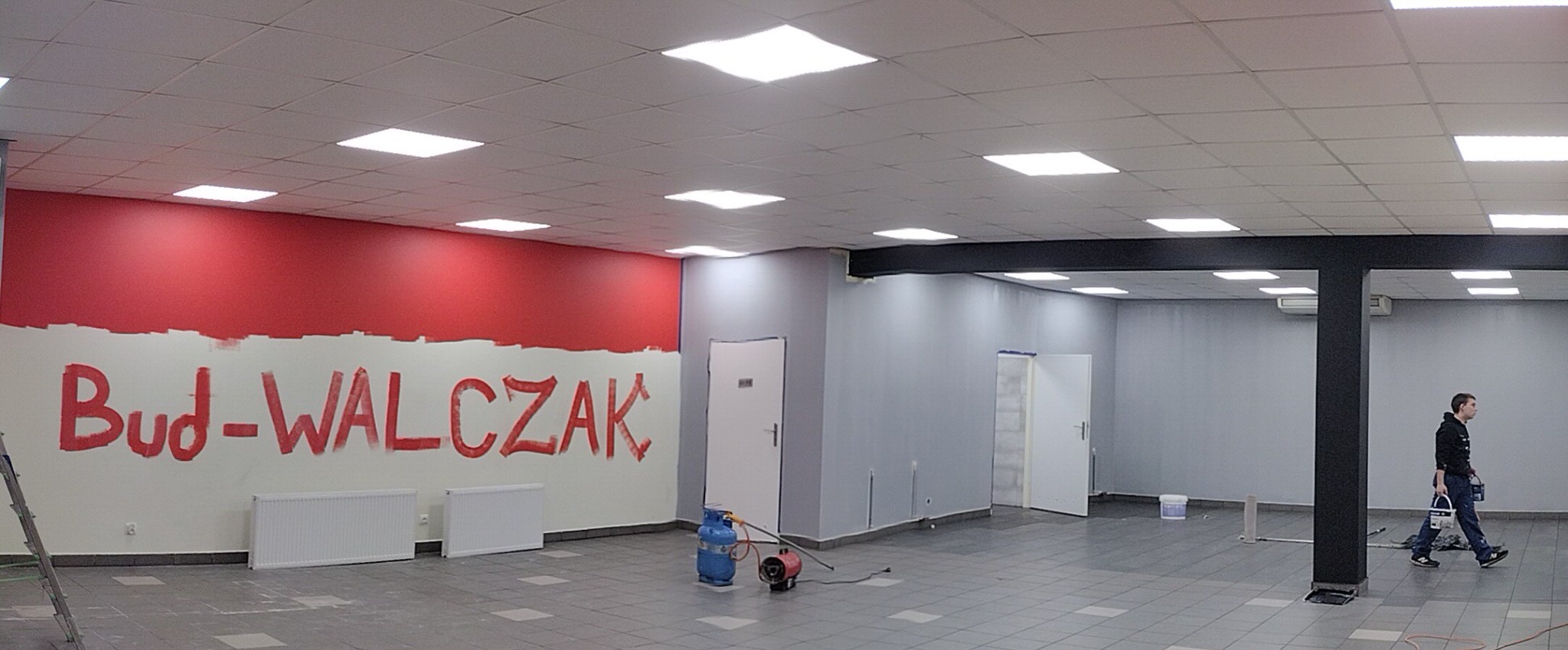 Wnętrze biura w trakcie malowania: część ściany pomalowana na czerwono z napisem 'Bud-WALCZAK', reszta ścian szara, widoczny grzejnik, drabina, nagrzewnica gazowa, oraz pracownik z wiadrem farby.