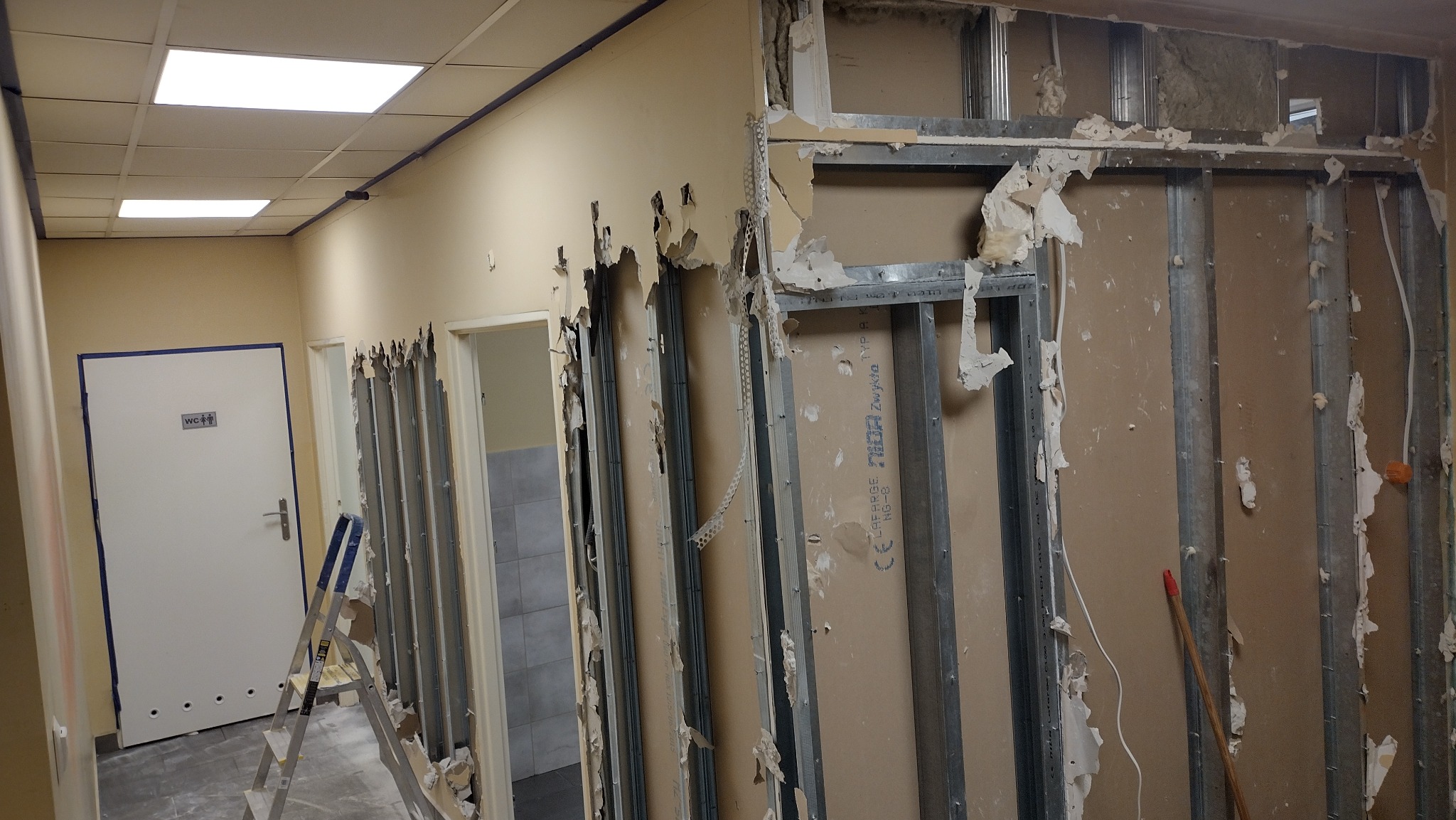 Wnętrze w trakcie demontażu ścian działowych, widoczne metalowe profile, fragmenty płyt kartonowo-gipsowych, instalacje elektryczne i drabina w korytarzu z drzwiami do toalety.