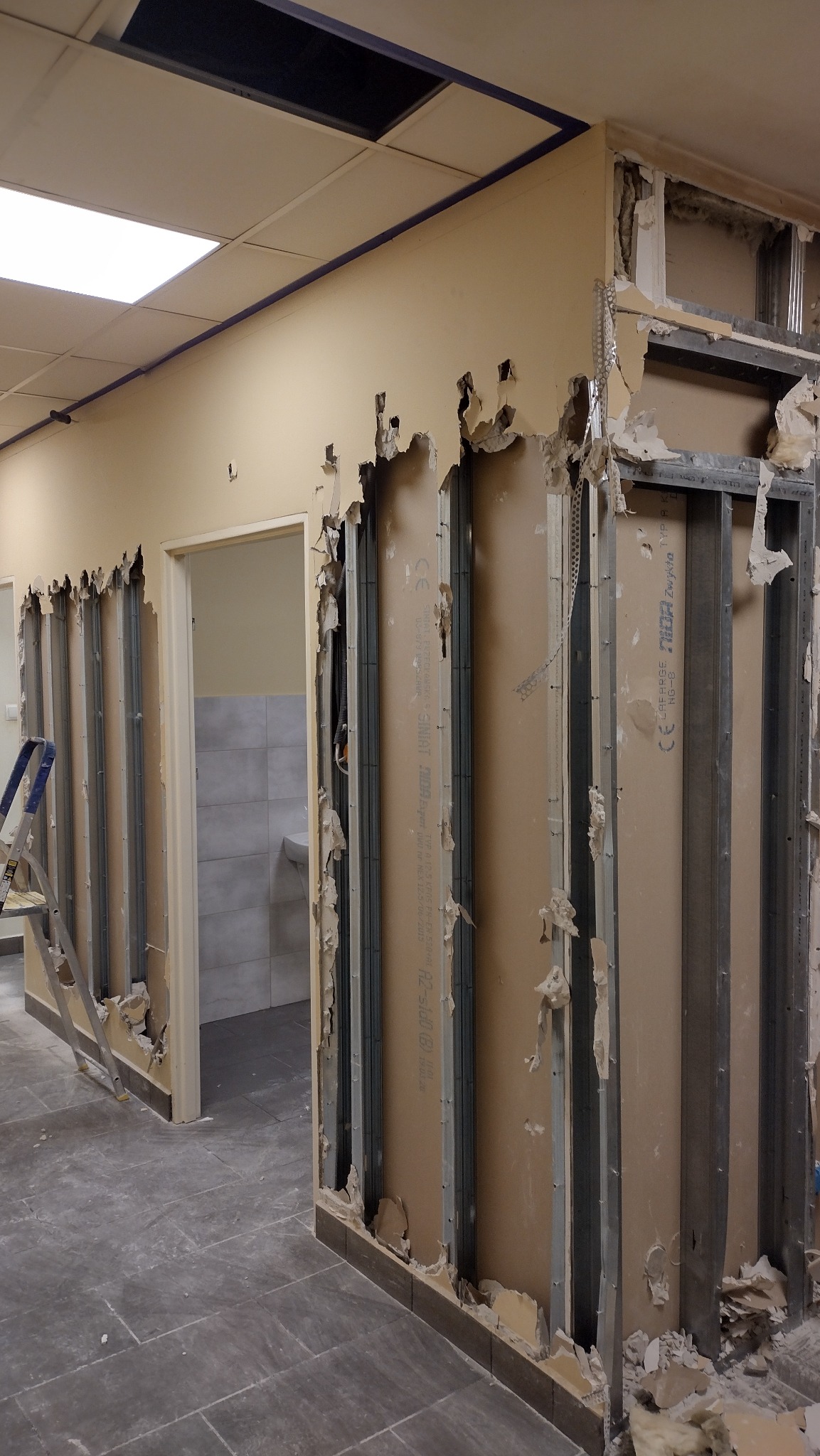 Demontaż ściany działowej z płyty kartonowo-gipsowej, widoczne metalowe profile i resztki płyty, w tle fragment łazienki z białymi płytkami.
