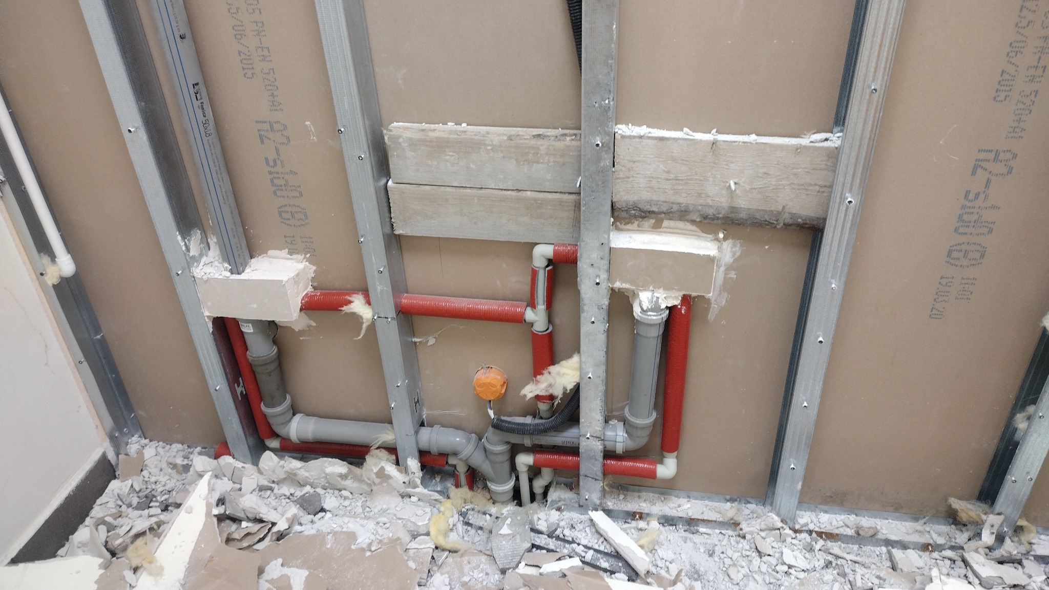 Instalacja wodno-kanalizacyjna w trakcie montażu, widoczne czerwone i szare rury, metalowe profile konstrukcyjne, płyty kartonowo-gipsowe oraz gruz na podłodze.