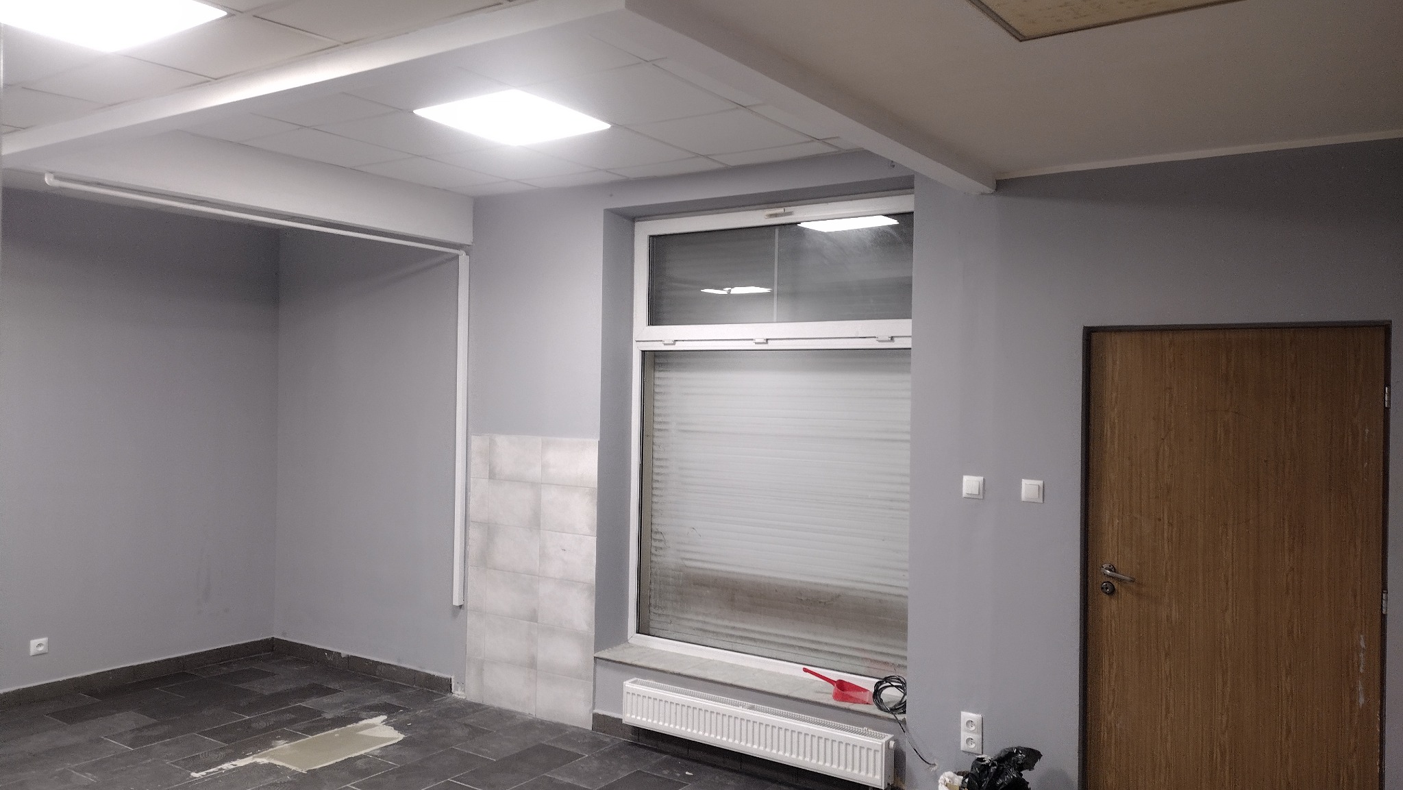 Pomieszczenie biurowe w trakcie remontu: szare ściany, okno z żaluzjami, grzejnik, częściowo położone płytki na ścianie, widoczne kable i narzędzia, podłoga z ciemnych płytek.