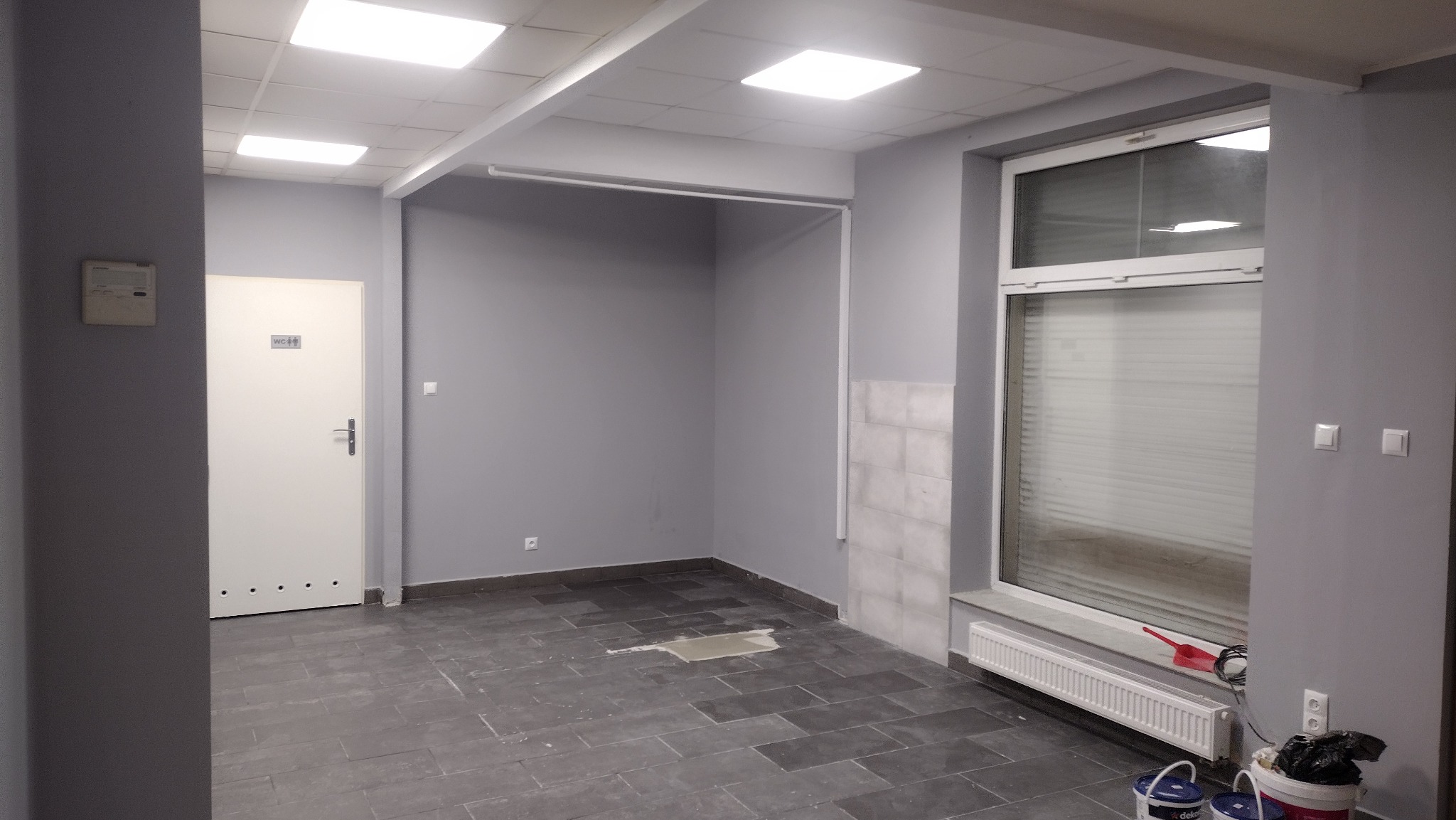 Szare pomieszczenie biurowe w trakcie prac wykończeniowych: widoczne szare ściany, płytki podłogowe, grzejnik pod oknem z roletami oraz wiaderka z farbą w rogu.
