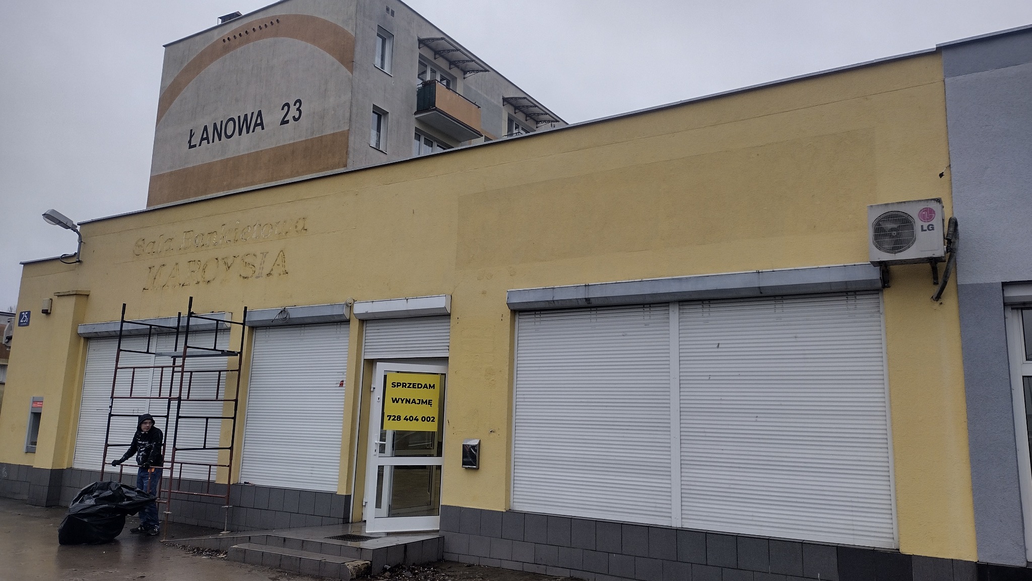 Żółta elewacja budynku z częściowo zdemontowanymi rusztowaniami, widoczne stare napisy i ogłoszenie o sprzedaży/wynajmie lokalu, osoba sprzątająca teren przed budynkiem.