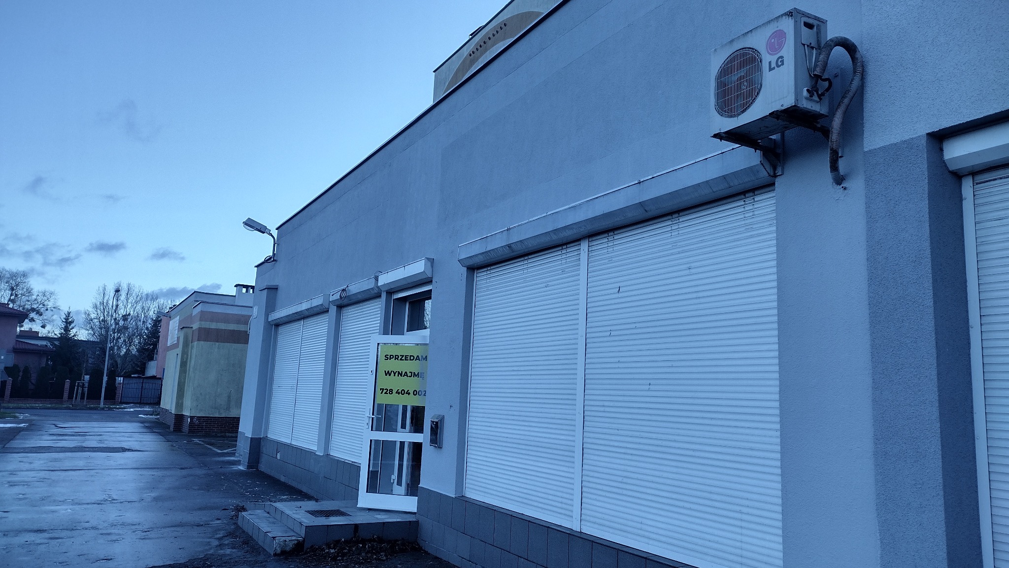Szary budynek usługowy z zamkniętymi roletami antywłamaniowymi, klimatyzator LG na elewacji i ogłoszeniem 'Sprzedam/Wynajmę' w drzwiach wejściowych.