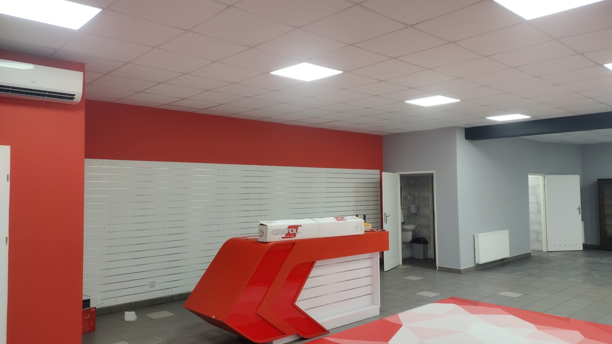 Wnętrze biura z pomalowanymi na czerwono ścianami i białymi panelami ściennymi, widoczna recepcja z opakowaniem folii Oracal, sufit z kwadratowymi lampami.