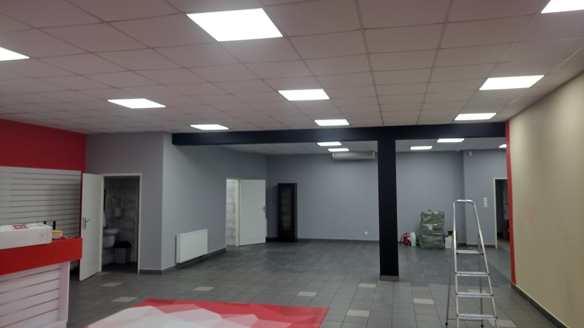 Przestronne wnętrze biurowe w trakcie odświeżania, z elementami czerwieni i szarości, widoczna drabina i przygotowane materiały budowlane, jasne oświetlenie sufitowe.