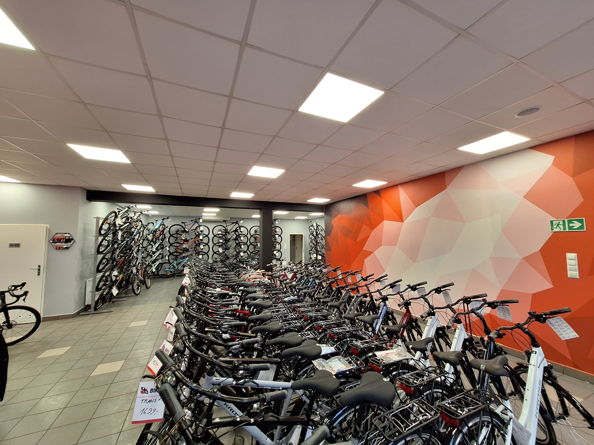 Rzędy rowerów w sklepie z pomarańczową ścianą w geometryczne wzory. Widoczny sufit kasetonowy z oświetleniem. Perspektywa z głębi sklepu.