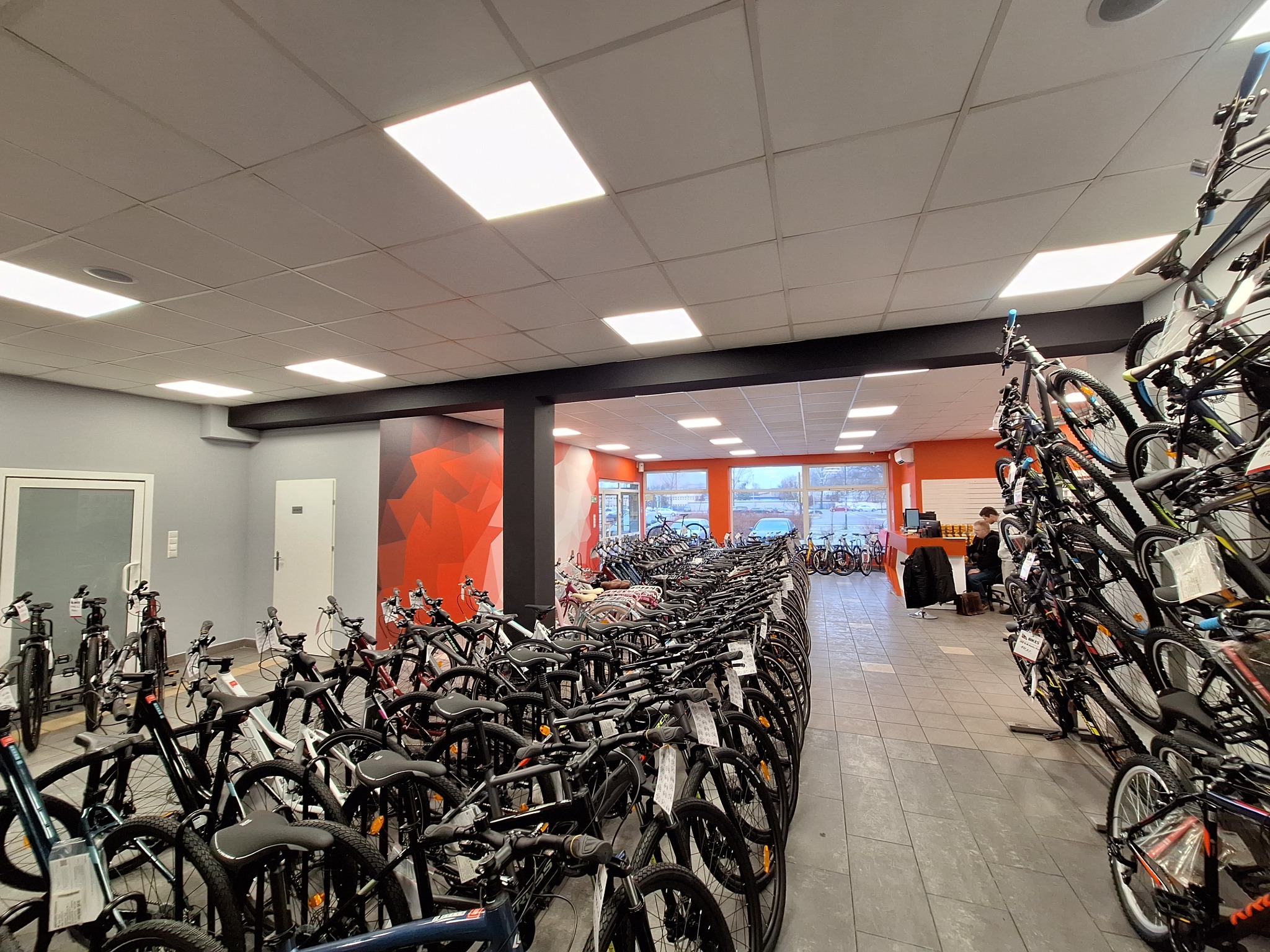 Wnętrze sklepu rowerowego z rzędami rowerów górskich i miejskich, widoczne białe ściany, sufit kasetonowy z oświetleniem LED, oraz pomarańczowy akcent kolorystyczny w głębi sklepu.