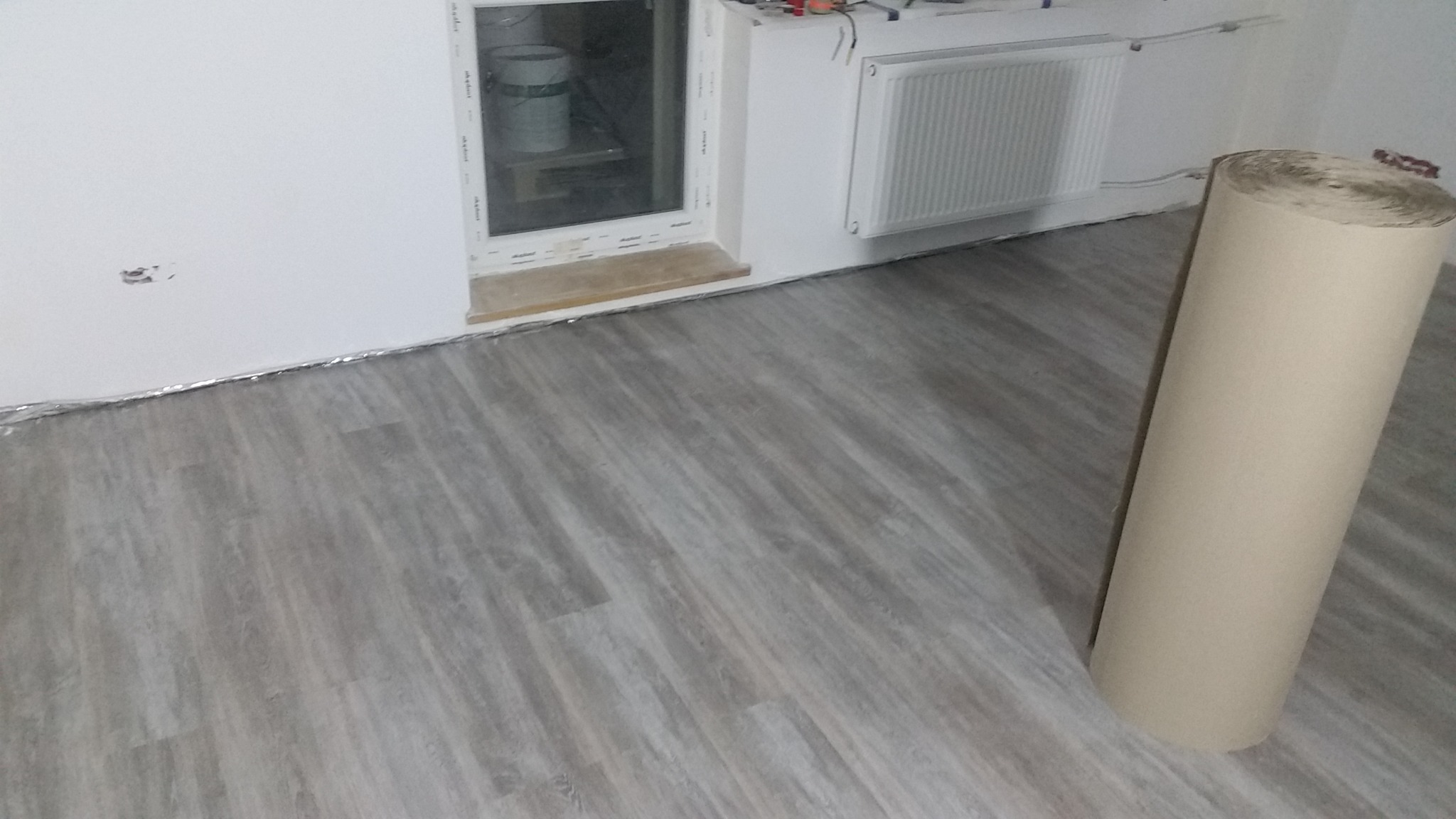 Jasnoszare panele podłogowe ułożone w pokoju, widoczna folia izolacyjna przy ścianie, okno z parapetem i grzejnik, obok stoi rolka papieru zabezpieczającego podłogę.