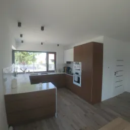 Jasne wnętrze kuchni z brązowymi i białymi szafkami, wyspą kuchenną z białym blatem, dużym oknem i drewnopodobną podłogą.