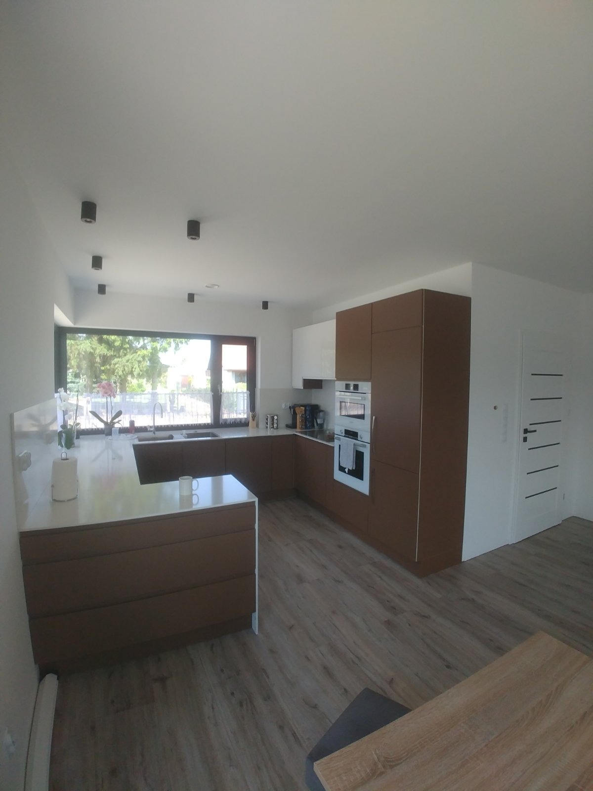 Jasne wnętrze kuchni z brązowymi i białymi szafkami, wyspą kuchenną z białym blatem, dużym oknem i drewnopodobną podłogą.