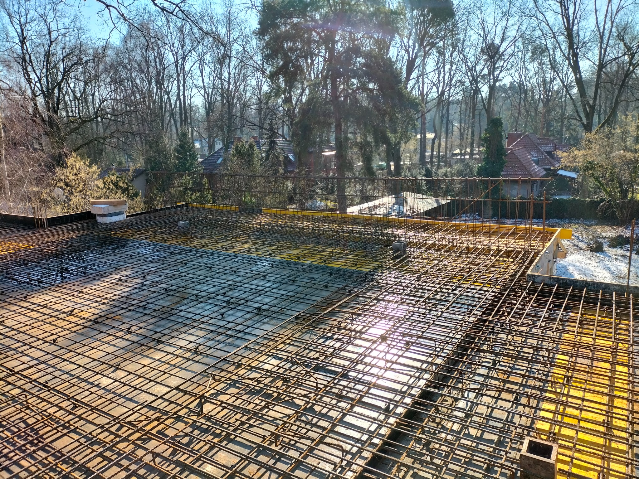 Zbrojenie stropu domu jednorodzinnego w trakcie budowy, widok z góry na siatkę z prętów stalowych, w tle drzewa bez liści i zabudowa jednorodzinna.