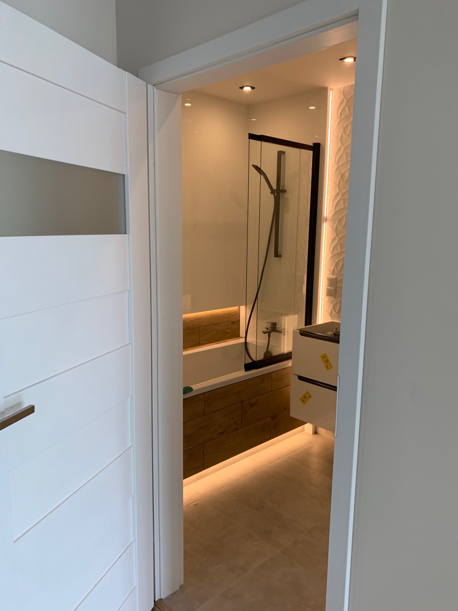 Wnętrze wykończonej łazienki z wanną obudowaną drewnopodobnymi płytkami, kabiną prysznicową z czarnymi ramami, białą umywalką z szufladami, oświetleniem LED i dekoracyjną ścianą z białych płytek 3D...