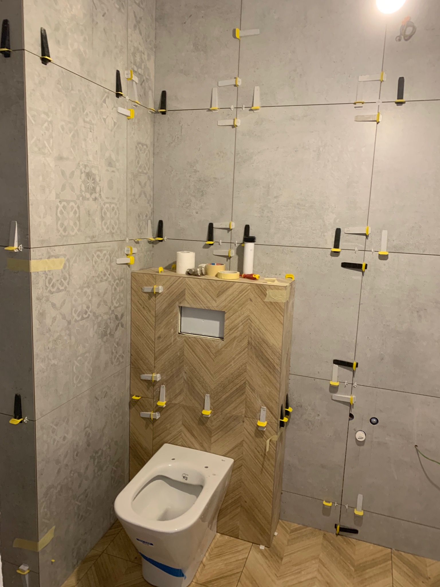 Łazienka w trakcie układania płytek: widoczne płytki ścienne imitujące beton z dekoracyjnym wzorem na jednej ścianie, płytki na obudowie stelaża WC ułożone w jodełkę, biała miska WC z niebieską...