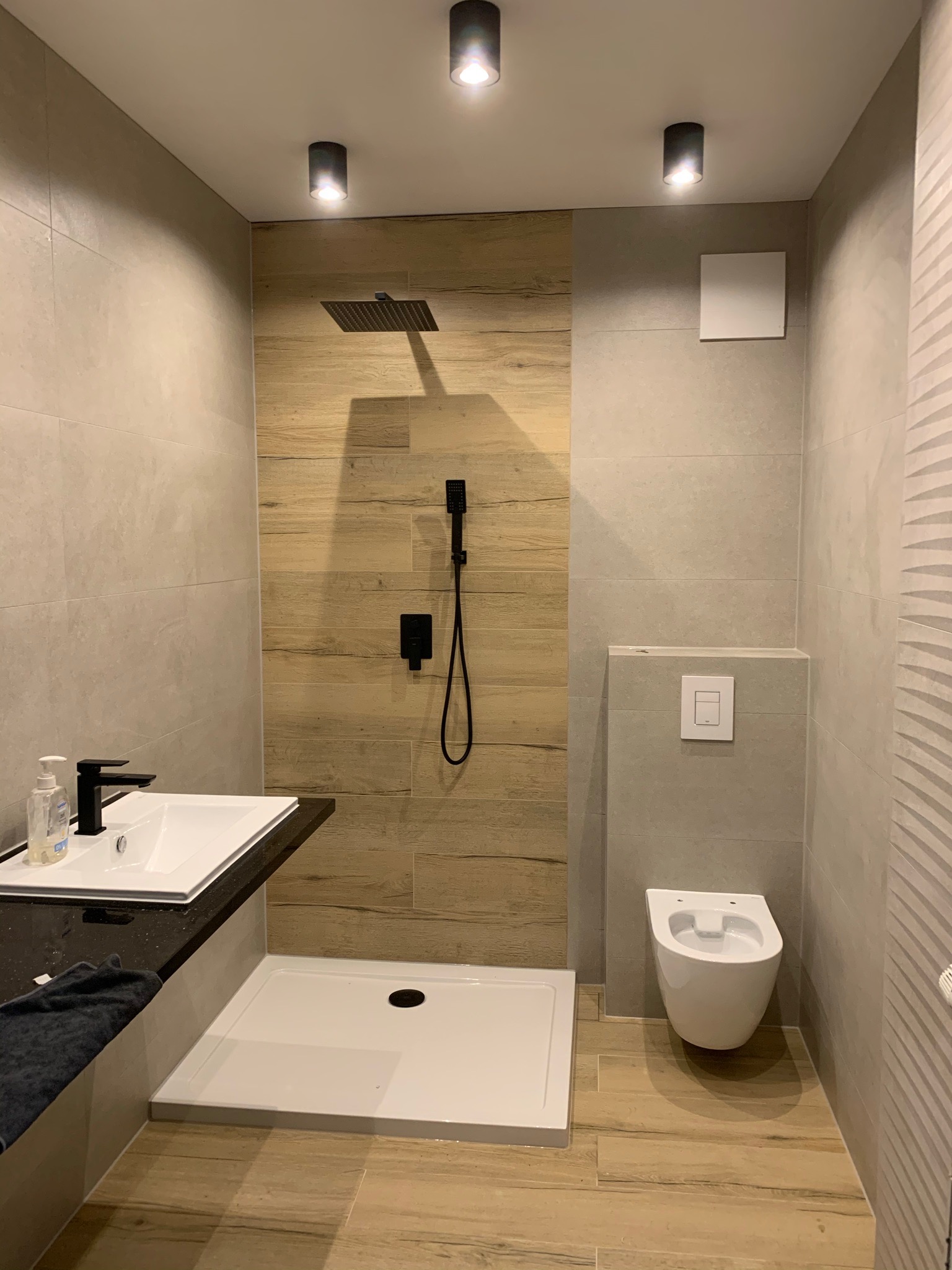 Nowoczesna łazienka z kabiną prysznicową typu walk-in, umywalką nablatową z czarnym blatem, wiszącą toaletą i drewnopodobnymi płytkami na ścianie pod prysznicem.