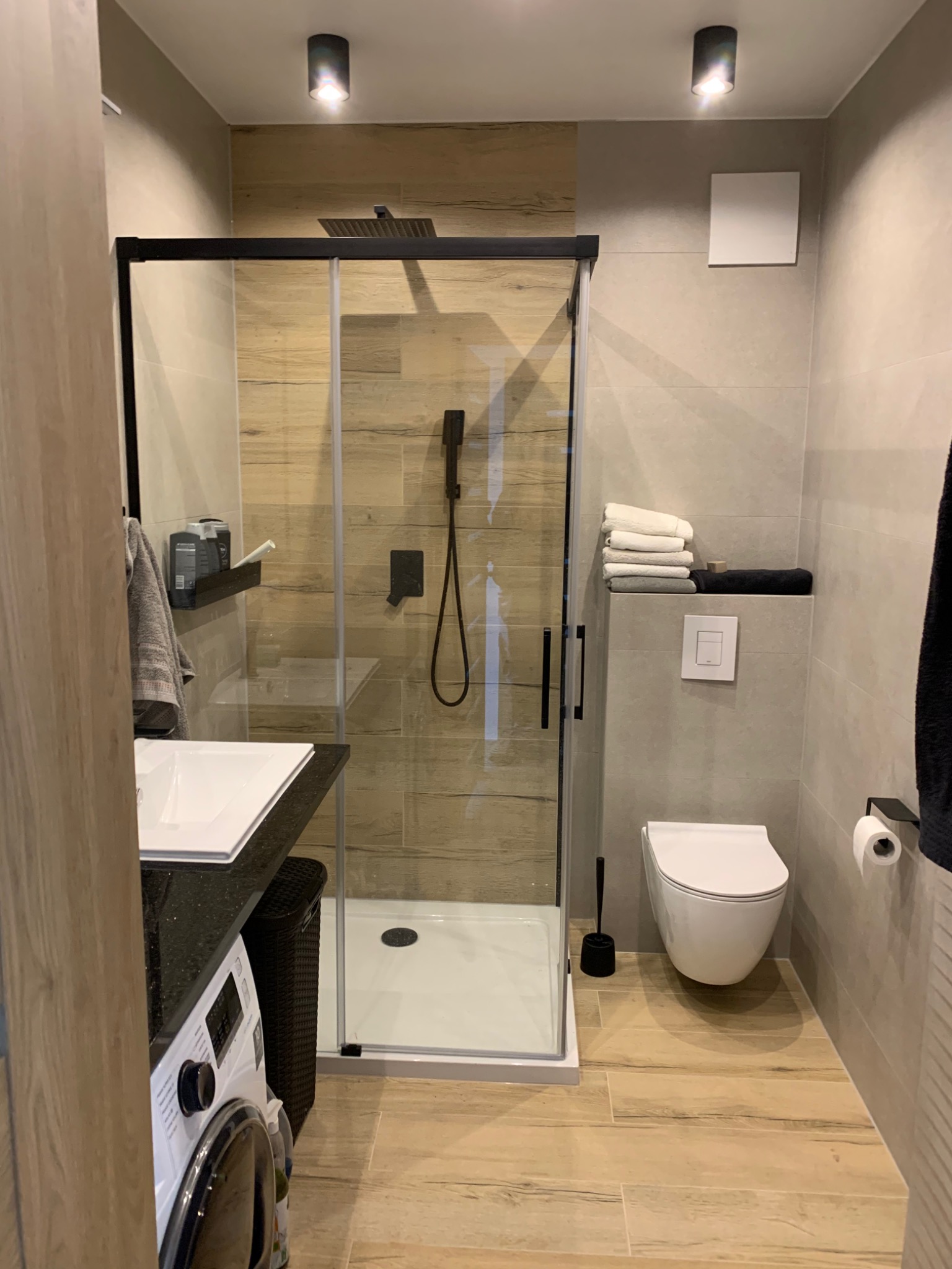 Nowoczesna łazienka w Krakowie z kabiną prysznicową typu walk-in z czarnymi elementami, umywalką z czarnym blatem, podwieszaną toaletą i pralką w tle.