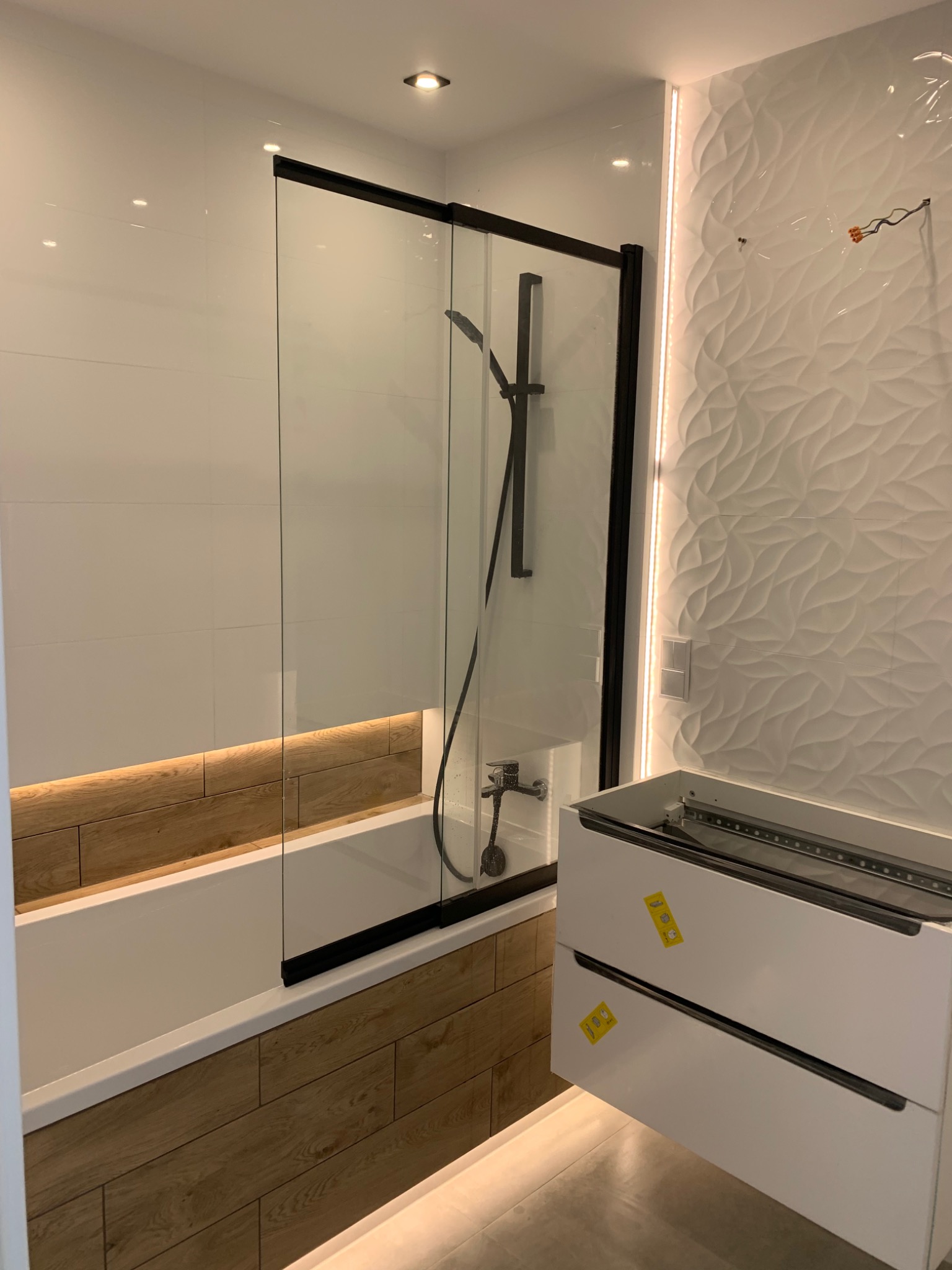 Nowoczesna łazienka z kabiną prysznicową typu walk-in z czarnymi profilami, obudową wanny imitującą drewno, dekoracyjną białą płytką 3D i podświetleniem LED przy ścianie. Częściowo zamontowana...