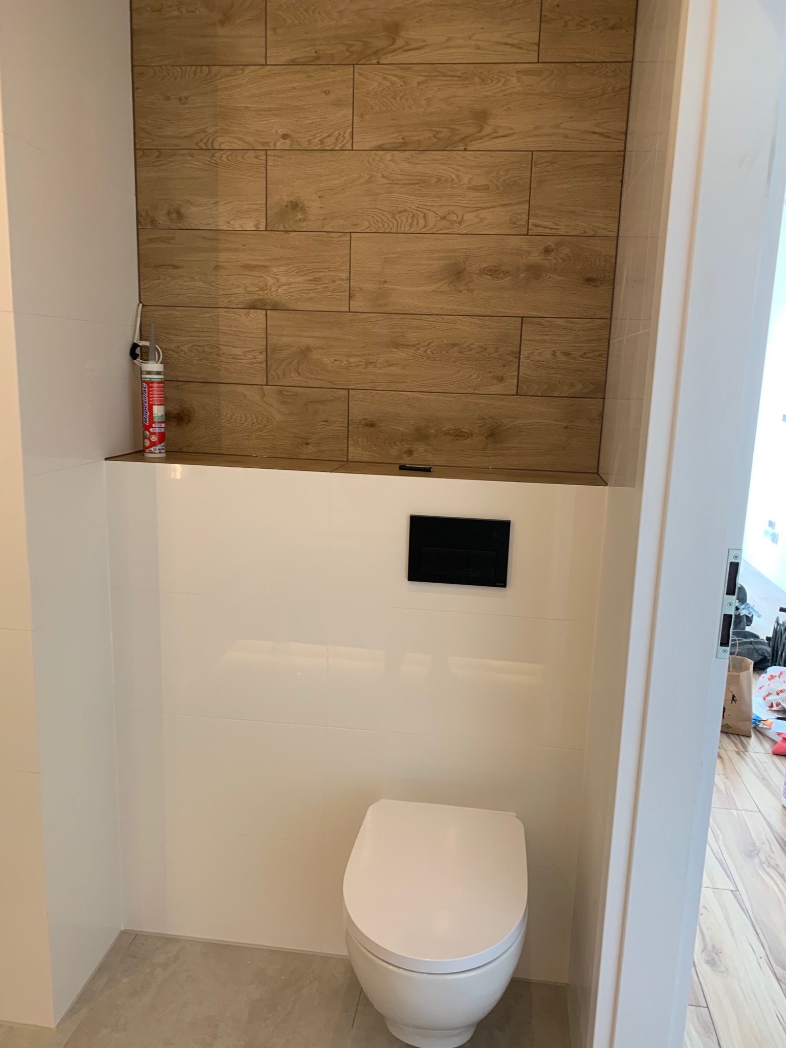 Nowoczesna toaleta w trakcie wykańczania: białe płytki ścienne, drewnopodobna okładzina nad stelażem WC, widoczna puszka z pianką montażową.