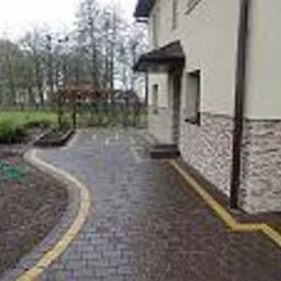 Ułożona kostka brukowa w kolorze grafitowym z żółtymi obrzeżami tworzy ścieżkę w ogrodzie przy domu z elewacją wykończoną kamieniem dekoracyjnym.