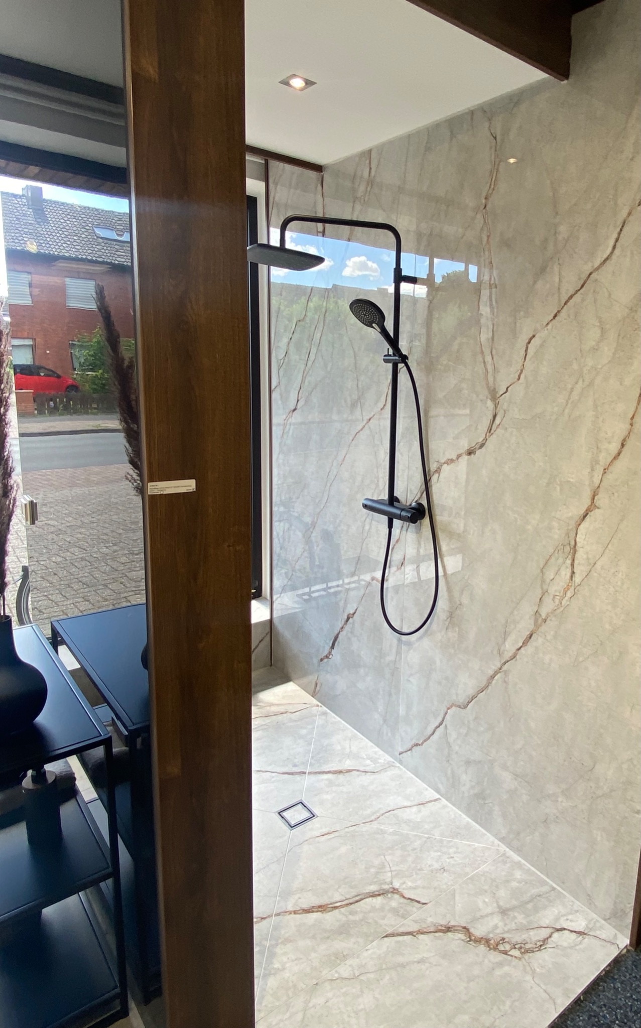 Wykończona łazienka z kabiną prysznicową typu walk-in, wyłożona płytkami imitującymi marmur z czarną armaturą, widok z zewnątrz przez wystawę sklepową.