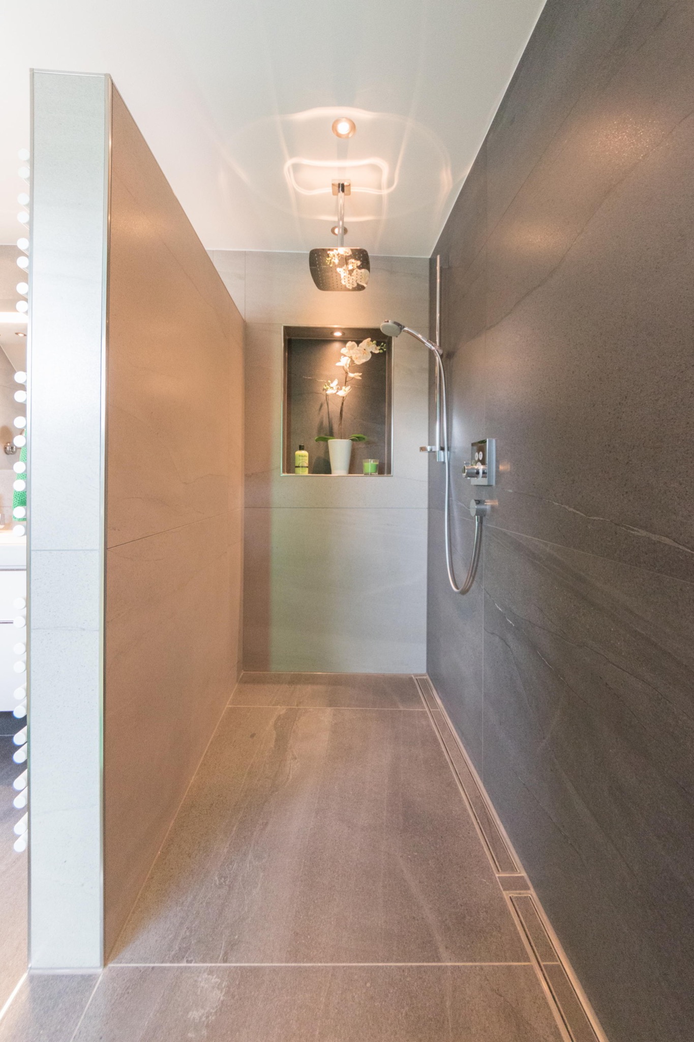 Nowoczesna kabina prysznicowa walk-in z szarymi płytkami imitującymi kamień, oświetloną wnęką z dekoracjami i odpływem liniowym w podłodze.