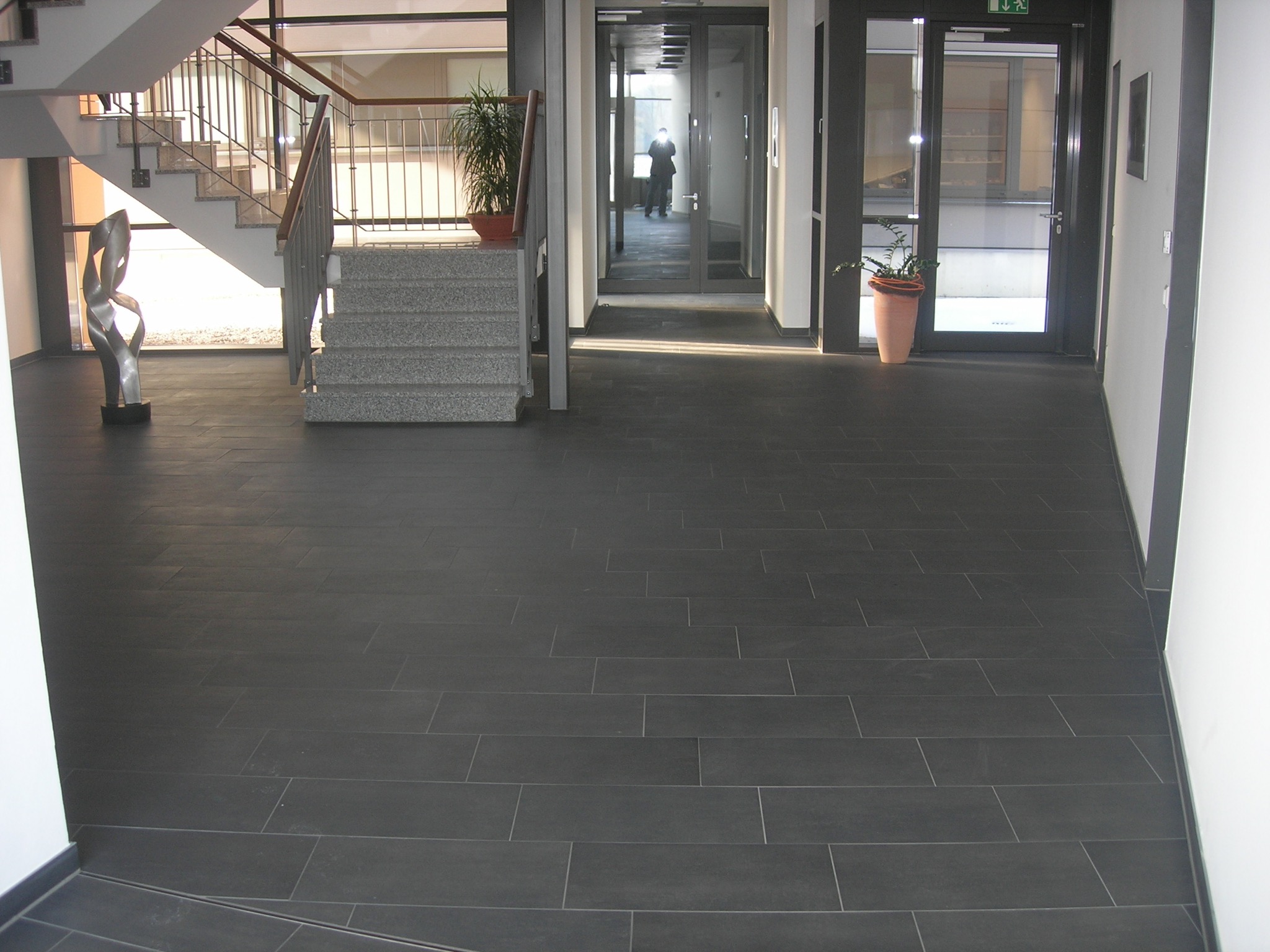 Ciemnoszara podłoga z płytek ceramicznych w holu wejściowym, widoczne schody, donica z rośliną i osoba w oddali przechodząca przez szklane drzwi.