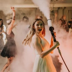 Fotografia Patrycja Wittke - Uśmiechnięta kobieta w białej sukience trzyma wyrzutnię dymu na parkiecie podczas imprezy, w tle tańczący goście i efekt mgły.