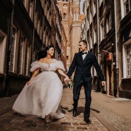 Fotografia Patrycja Wittke - Para młoda, trzymając się za ręce, biegnie po brukowanej uliczce Starego Miasta, w tle wysoka wieża kościelna.