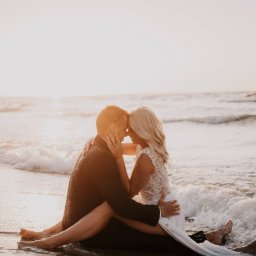 Fotografia Patrycja Wittke - Para młoda siedzi na plaży w objęciach, fale delikatnie obmywają ich stopy, a zachodzące słońce tworzy romantyczną aurę.