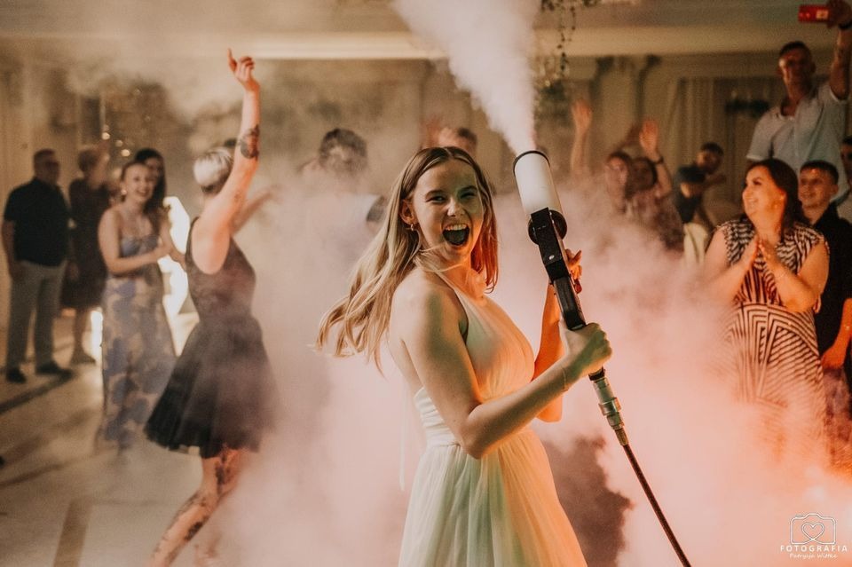 Uśmiechnięta kobieta w białej sukience trzyma wyrzutnię dymu na parkiecie podczas imprezy, w tle tańczący goście i efekt mgły.