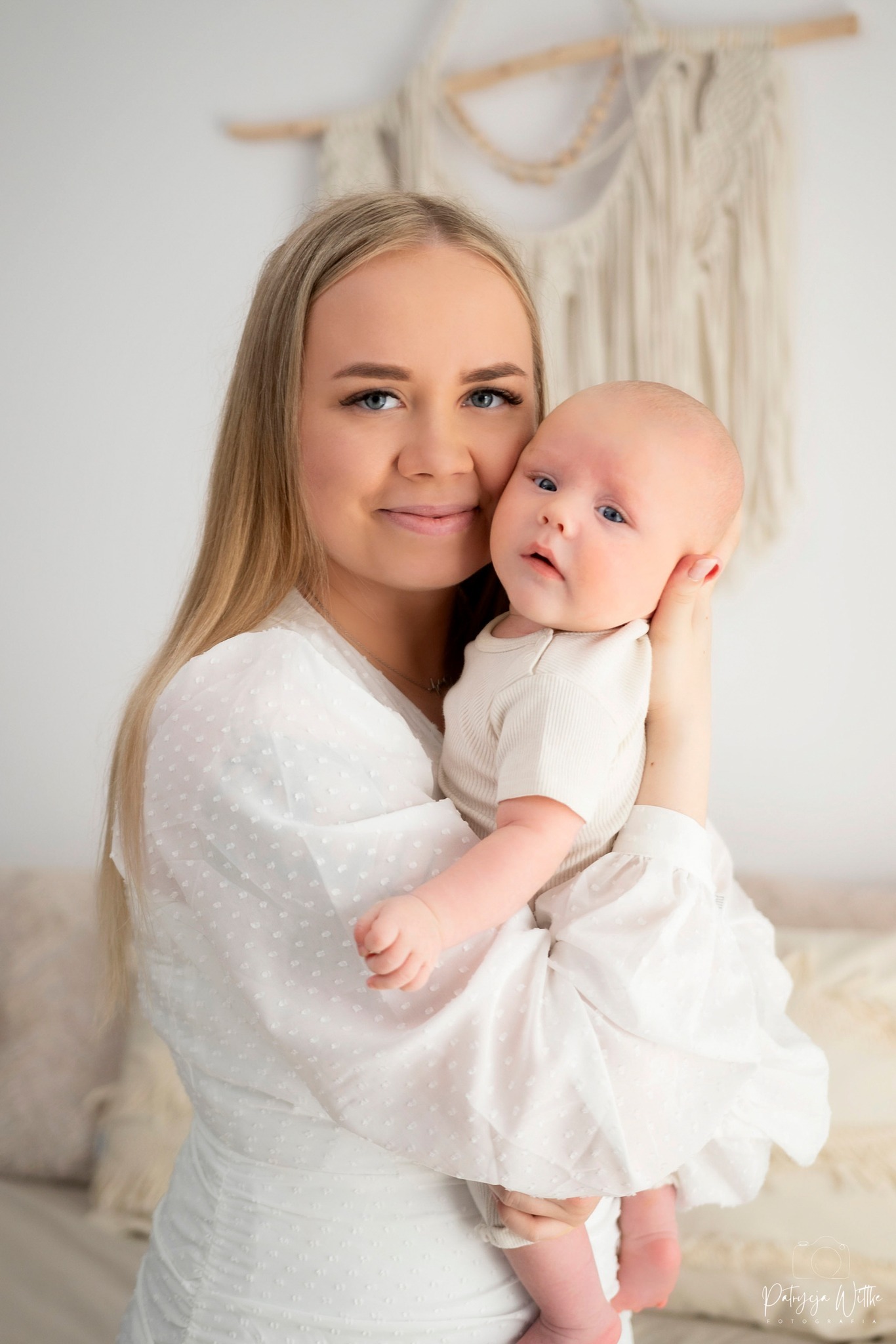 Uśmiechnięta blondynka trzyma na rękach niemowlę ubrane w białe ubranko, w tle dekoracyjny makramowy element na ścianie, delikatne oświetlenie.