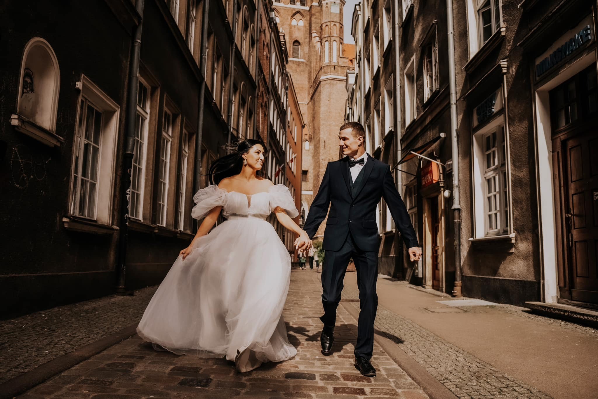 Para młoda, trzymając się za ręce, biegnie po brukowanej uliczce Starego Miasta, w tle wysoka wieża kościelna.