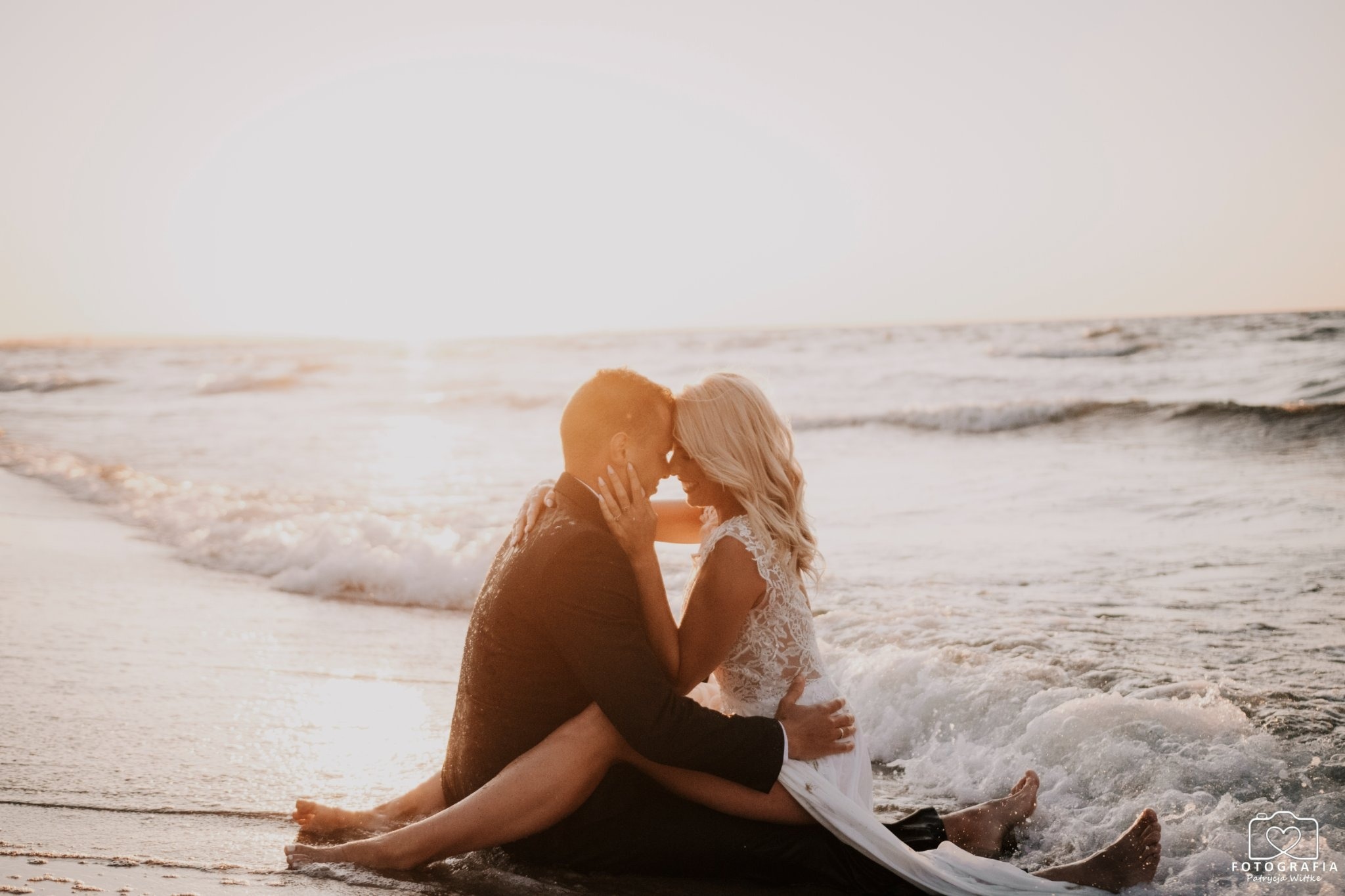 Para młoda siedzi na plaży w objęciach, fale delikatnie obmywają ich stopy, a zachodzące słońce tworzy romantyczną aurę.