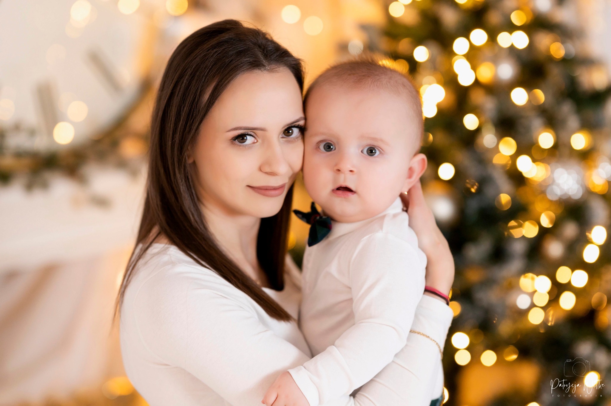 Uśmiechnięta mama tuli do siebie niemowlę z zieloną muchą na tle rozświetlonej choinki i dużego zegara.