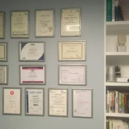 Ściana z szarym tłem, na której wiszą w kilku rzędach oprawione certyfikaty i dyplomy, obok biały regał z książkami, głośnikiem i projektorem.