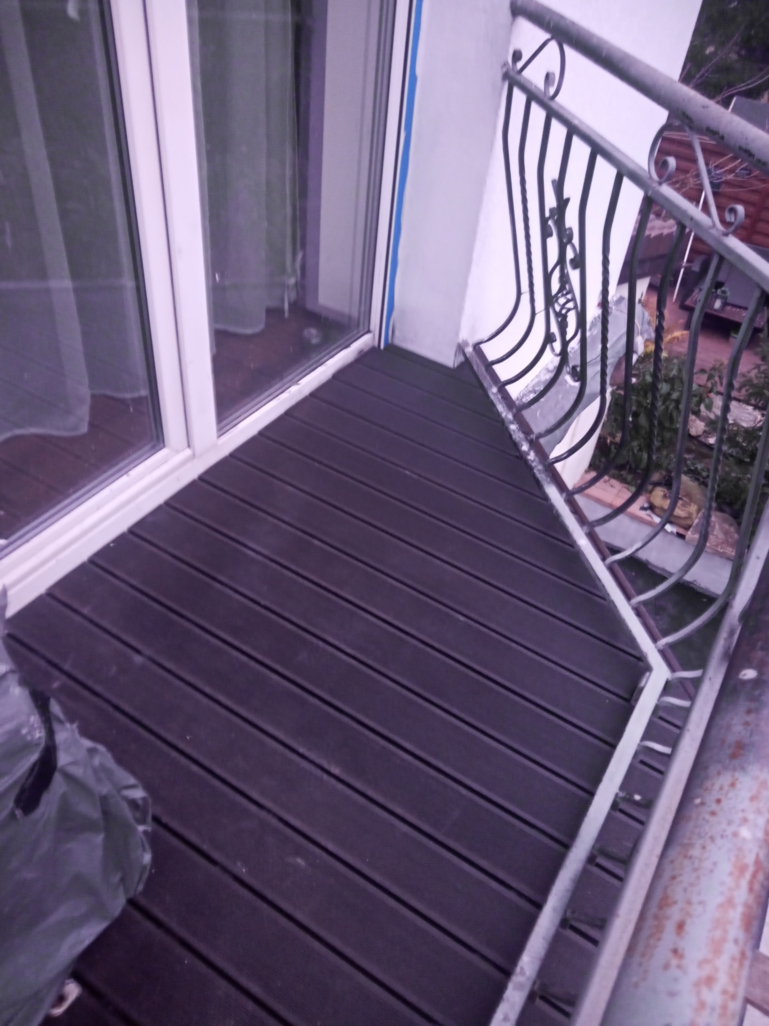 Balkon z nową, ciemną podłogą z desek kompozytowych, widok z góry na metalową barierkę i okno balkonowe.