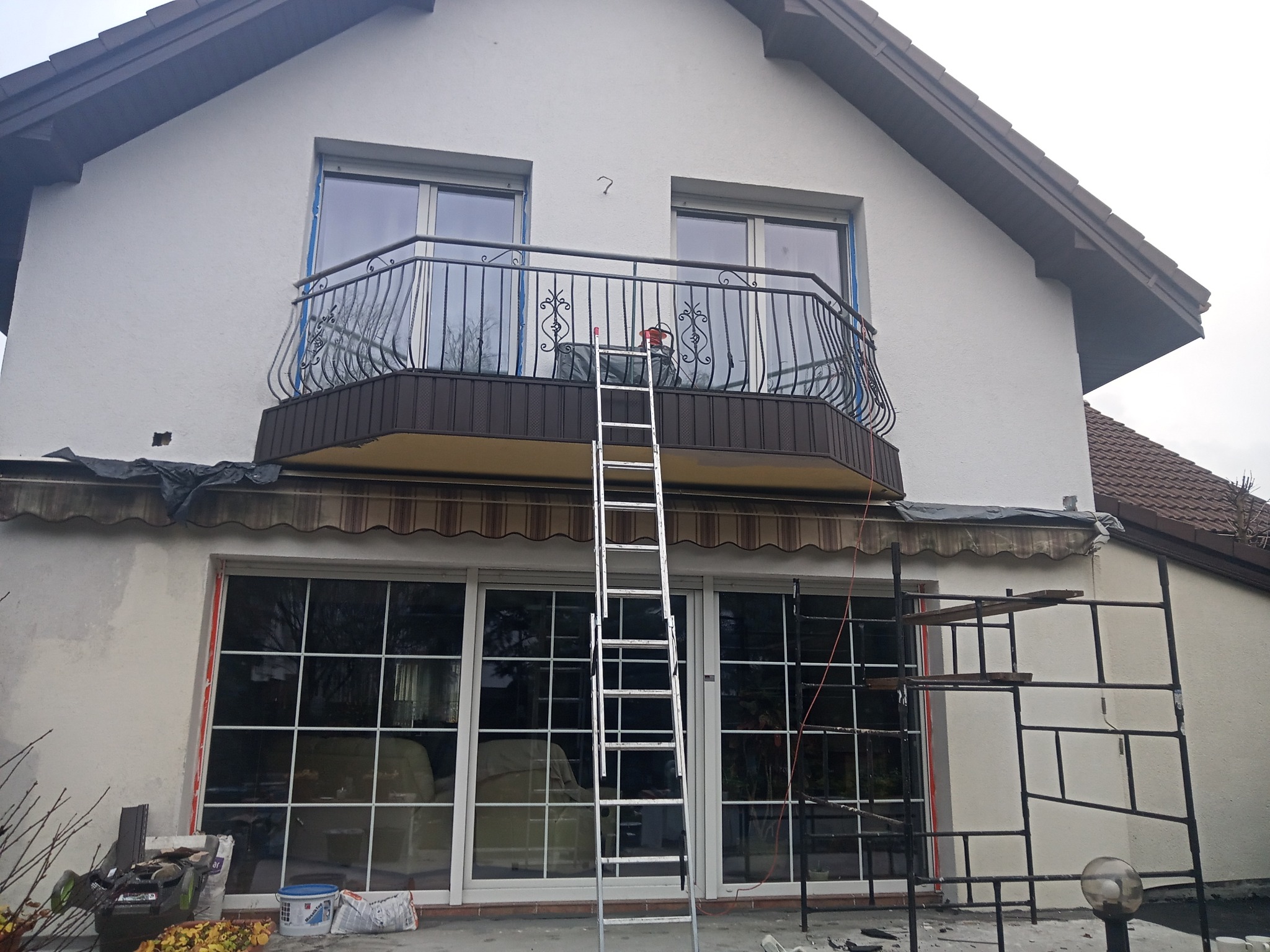 Dwukondygnacyjny dom z balkonem w trakcie przygotowań do malowania elewacji, widoczne zabezpieczone okna, rozstawiona drabina i rusztowanie, farby i narzędzia malarskie leżące na ziemi.