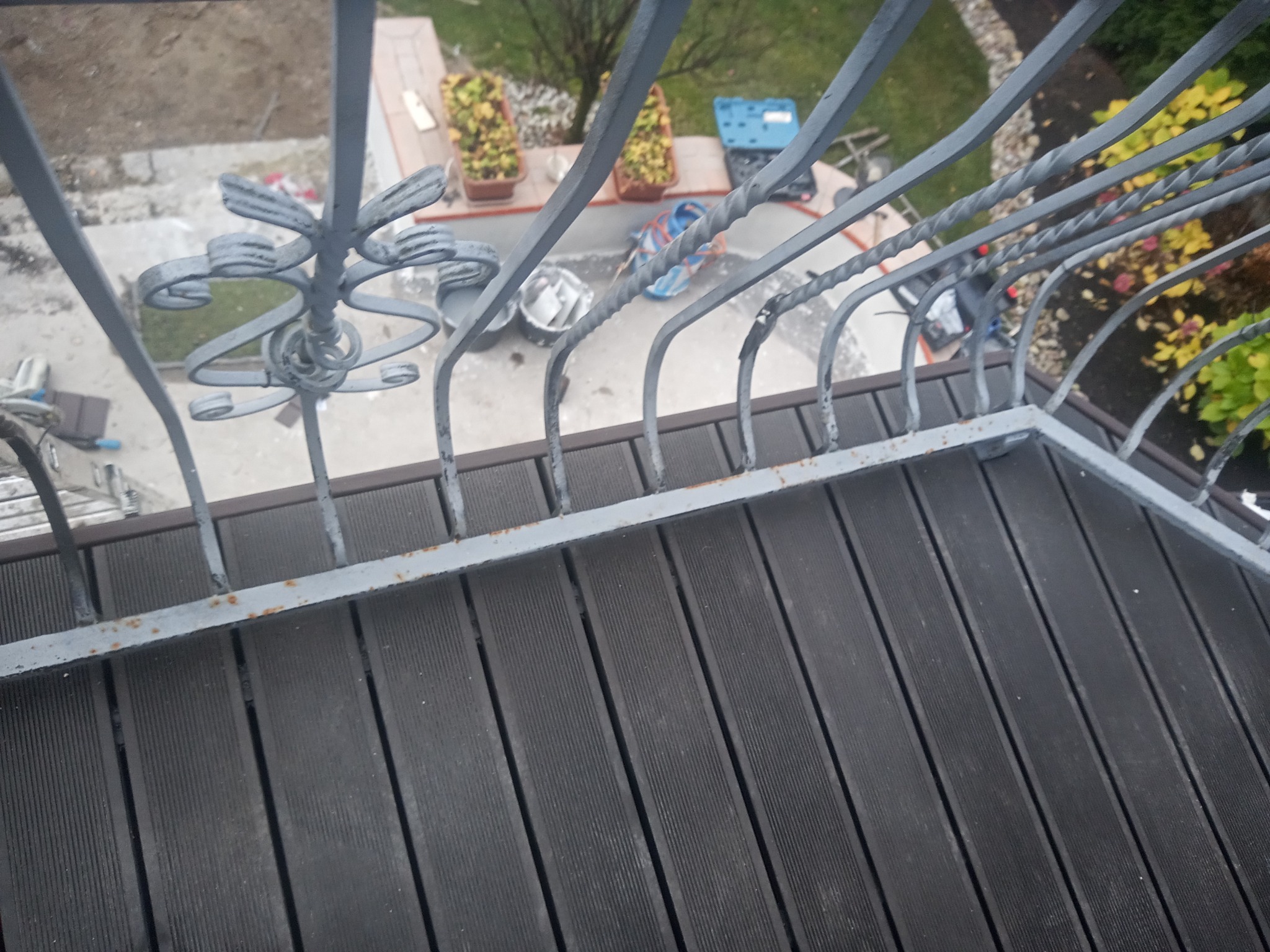 Perspektywa z góry na balkon z szarymi metalowymi balustradami z dekoracyjnymi elementami i ciemnobrązową podłogą z desek kompozytowych; widoczne narzędzia i doniczki na zewnątrz.