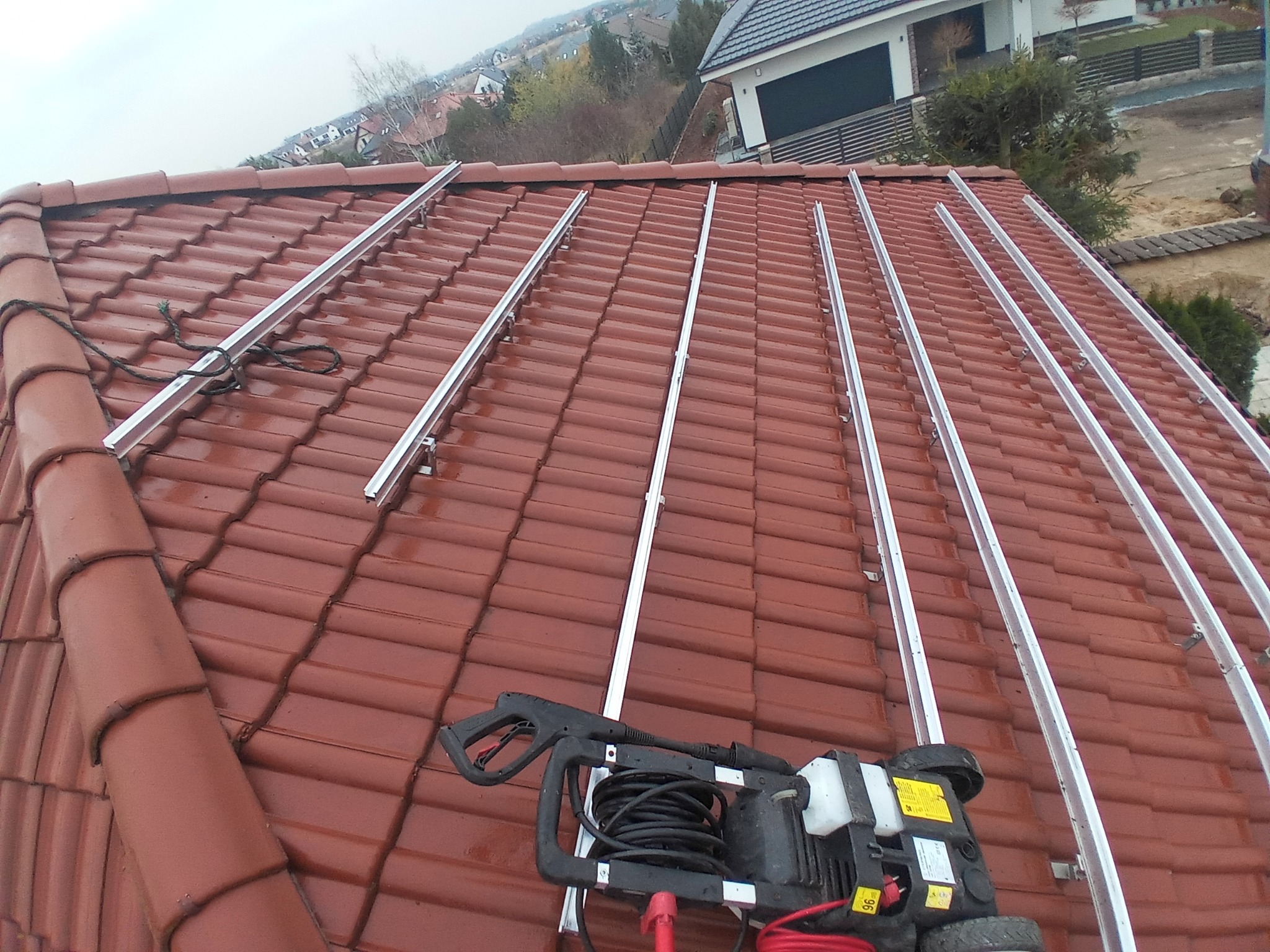 Czerwona dachówka z zamontowanymi aluminiowymi szynami i myjką ciśnieniową u dołu kadru, przygotowanie do montażu paneli fotowoltaicznych.