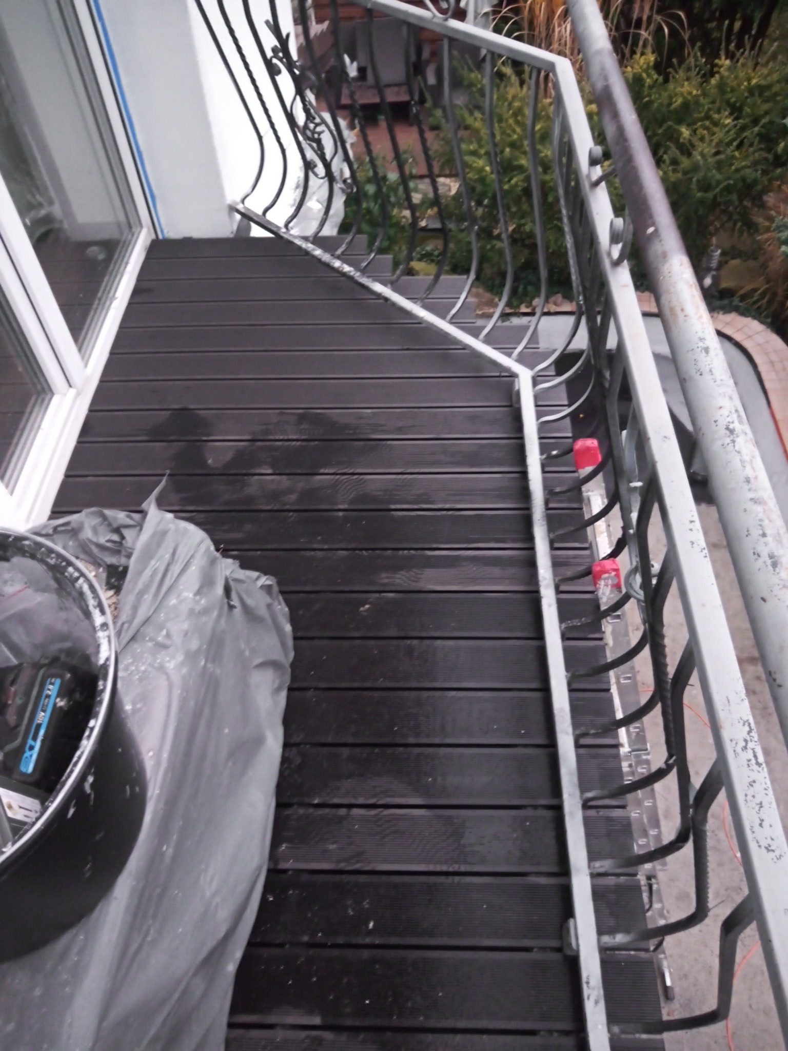 Ciemne deski tarasowe na balkonie po umyciu, widoczne ślady wilgoci, metalowa barierka z ozdobnymi elementami, przykryte folią wiadro z narzędziami.