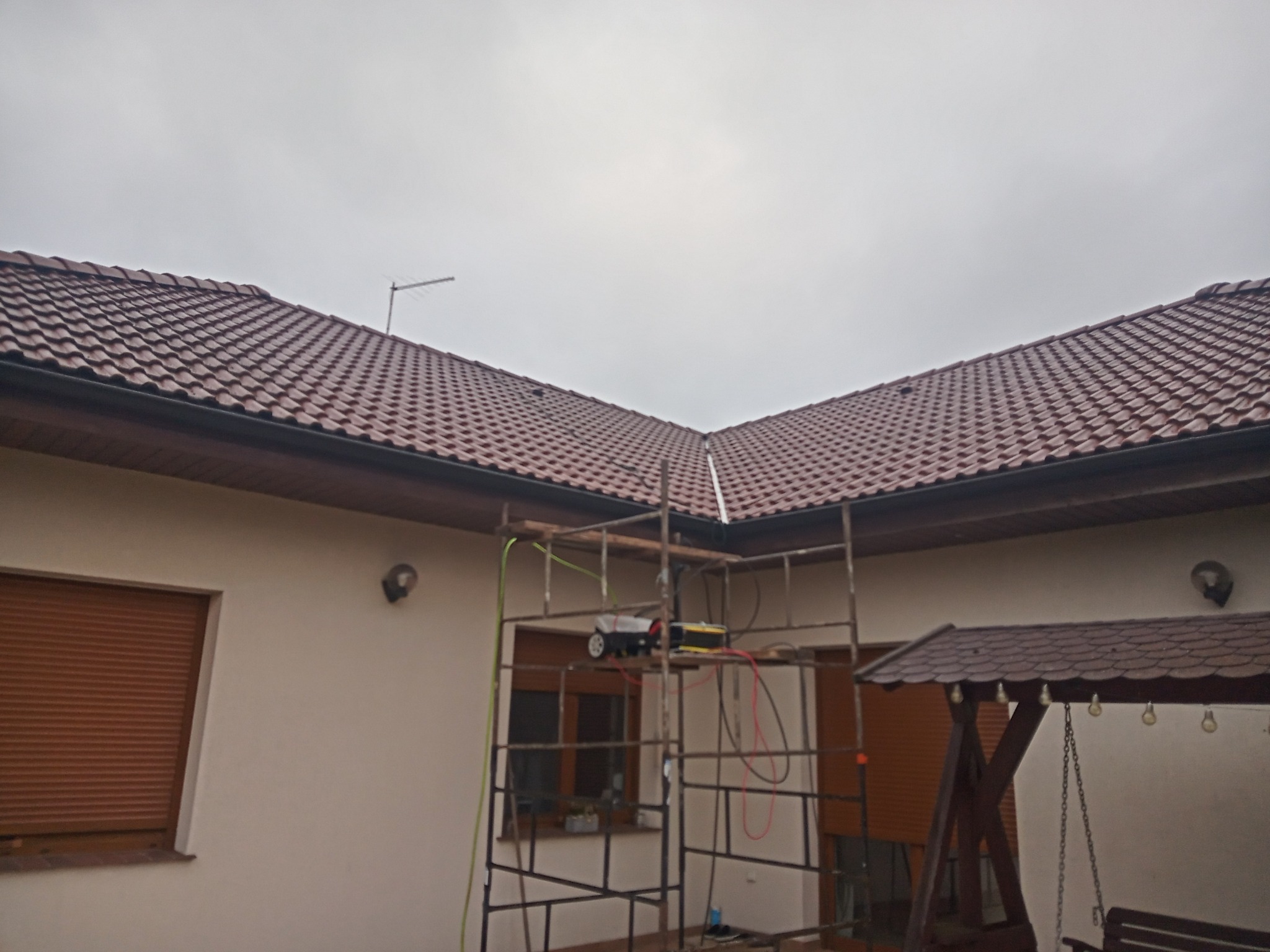 Metalowe rusztowanie ustawione przy ścianie domu z brązowym dachem z dachówki, na rusztowaniu leży sprzęt elektryczny, widoczne kable i wąż, pochmurne niebo w tle.