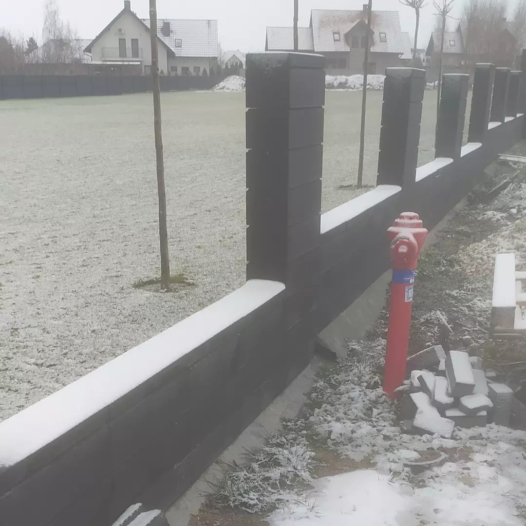 Ciemna, modułowa elewacja ogrodzenia z betonowych bloczków, lekko przyprószona śniegiem, z widocznym hydrantem przeciwpożarowym i stertą bloczków u podstawy, na tle zaśnieżonego trawnika i domów.